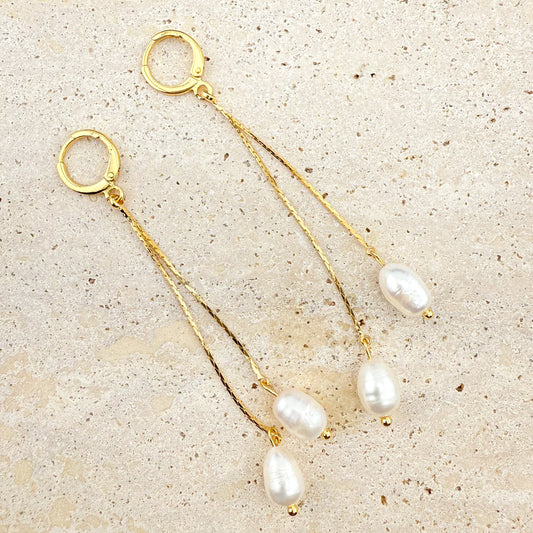 A pair of elegant Aking Sinta gold huggie hoop earrings, each featuring two delicate, dangling gold chains of varying lengths, finished with lustrous white pearls. The earrings are arranged on a light, textured natural stone background, highlighting the classic, sophisticated contrast between the polished gold and the radiant white pearls