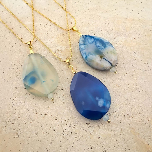 A collection of three elegant Aking Sinta gold chain necklaces, each featuring a unique, faceted teardrop-shaped agate pendant in varying shades of blue, white, and indigo. The necklaces are beautifully arranged on a light, textured natural stone surface, highlighting the vibrant natural patterns and semi-translucent quality of the agate gemstones