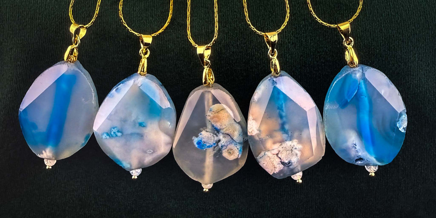 A stunning collection of five Aking Sinta gold chain necklaces, each featuring a faceted, teardrop-shaped blue agate pendant. The gemstones reveal a range of unique, ethereal patterns, from vibrant blue plumes to soft, earthy inclusions, elegantly arranged in a row against a solid black background to highlight their intricate textures and natural beauty