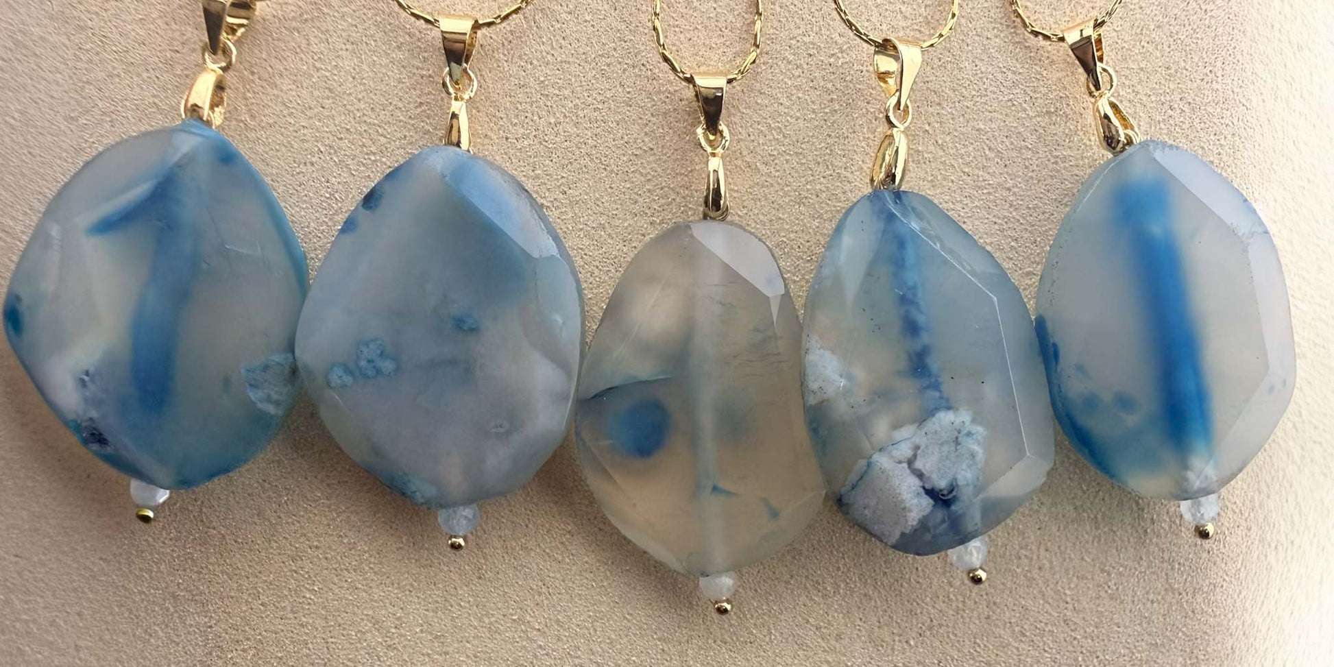 A detailed close-up of a collection of five Aking Sinta gold chain necklaces, each featuring a faceted teardrop-shaped blue agate pendant. The gemstones showcase unique, organic blue and white patterns, beautifully arranged against a neutral, textured display background to emphasize their natural stone beauty and craftsmanship