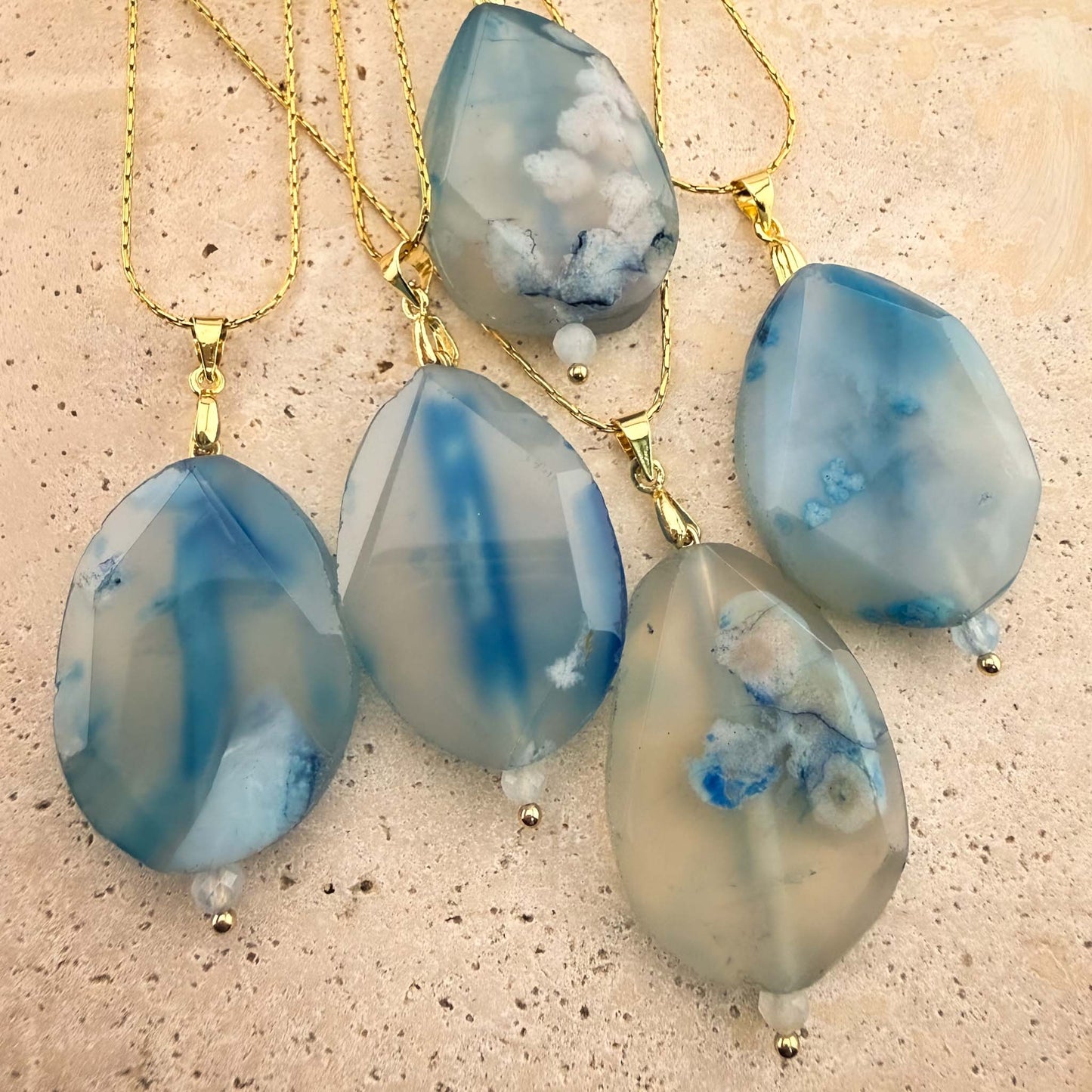 A stunning collection of five Aking Sinta gold chain necklaces, each featuring a faceted, teardrop-shaped blue agate pendant. The gemstones showcase a variety of unique, ethereal patterns, ranging from translucent white with blue plumes to bold, solid indigo bands, elegantly arranged on a light, textured natural stone surface