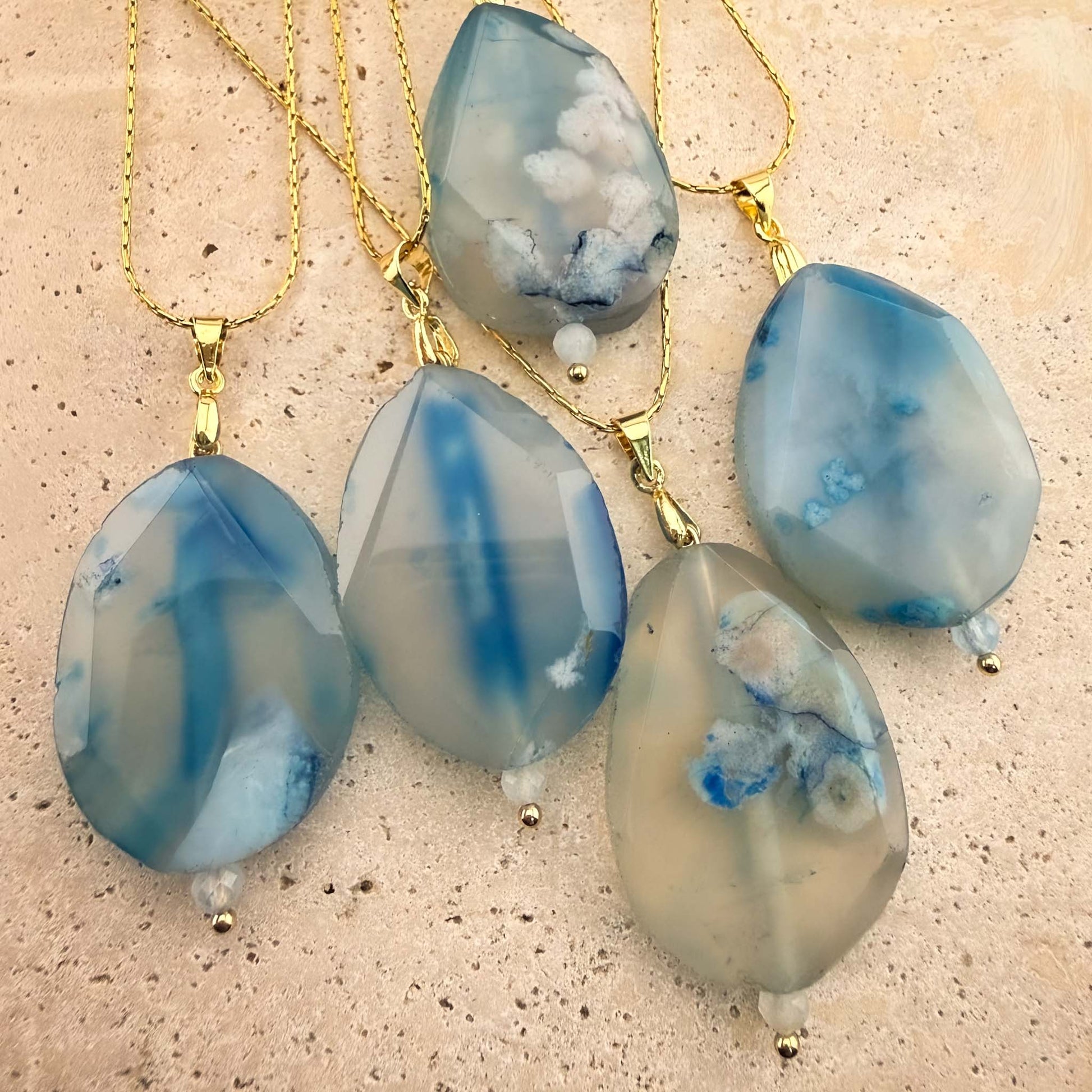 A stunning collection of five Aking Sinta gold chain necklaces, each featuring a faceted, teardrop-shaped blue agate pendant. The gemstones showcase a variety of unique, ethereal patterns, ranging from translucent white with blue plumes to bold, solid indigo bands, elegantly arranged on a light, textured natural stone surface