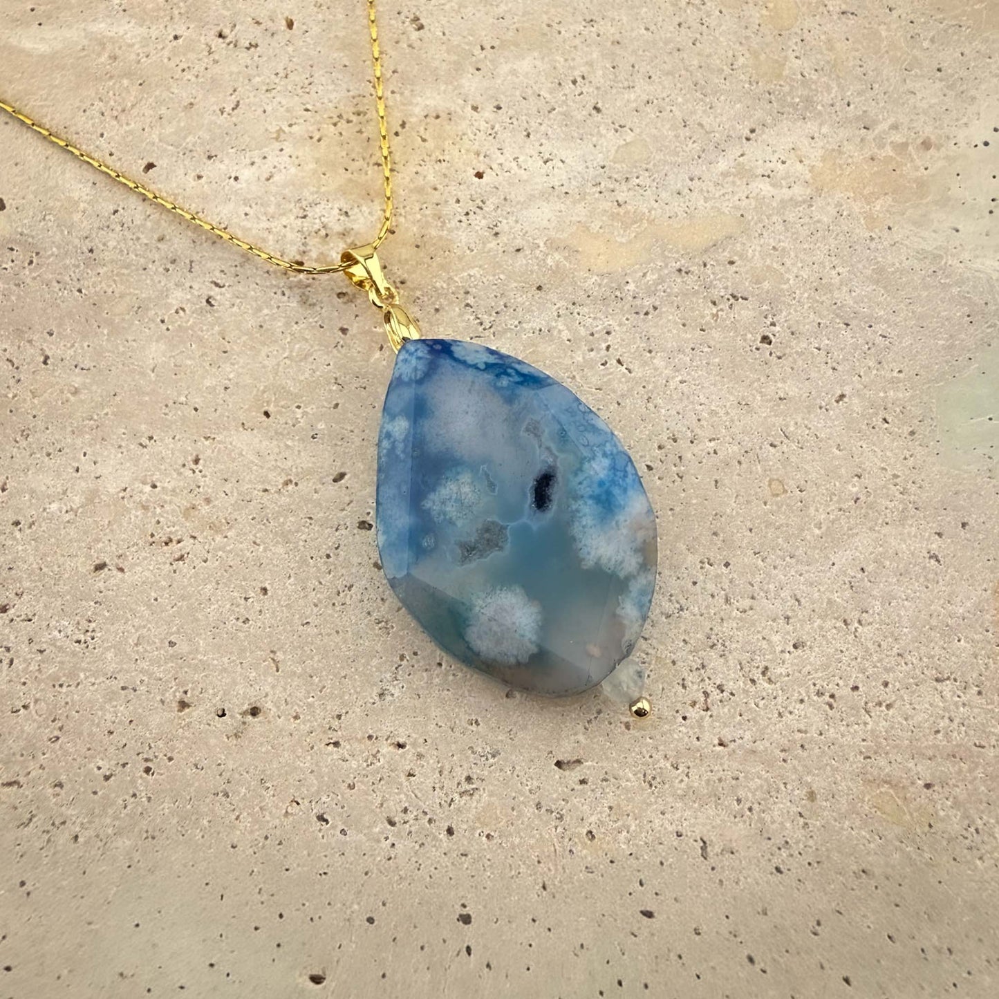 A detailed close-up of an Aking Sinta gold chain necklace featuring a faceted teardrop-shaped blue agate pendant. The gemstone displays intricate, cloud-like white inclusions against a soft blue backdrop, elegantly presented on a light, textured natural stone surface