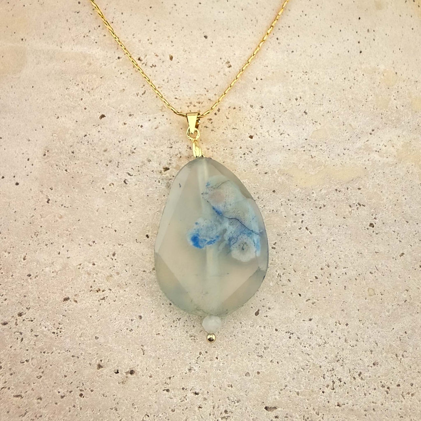 A close-up view of an Aking Sinta gold chain necklace featuring a faceted, translucent teardrop-shaped agate pendant. The stone exhibits ethereal white and soft blue plume inclusions, elegantly showcased against a light, textured natural stone surfaceon a marble surface.
