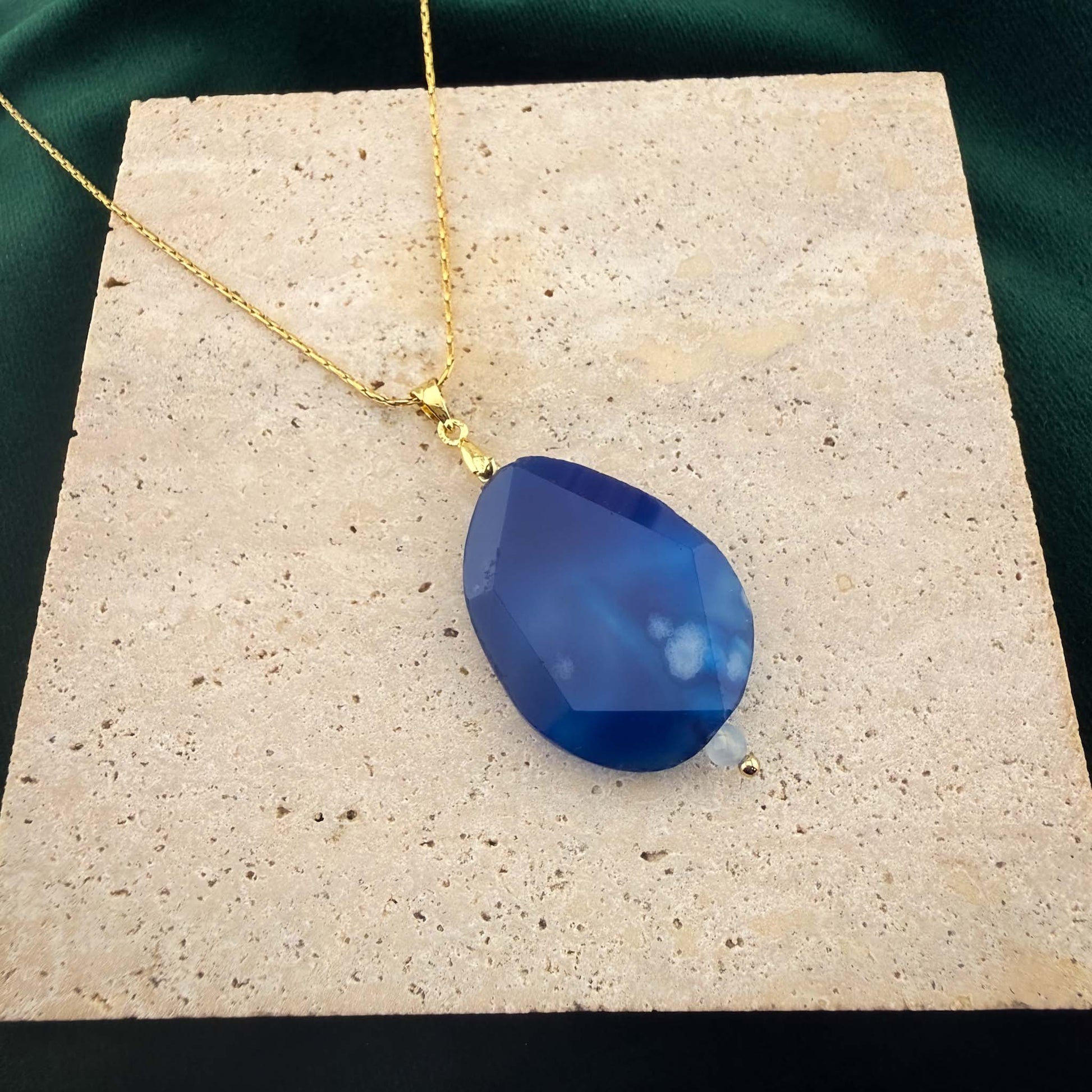 A detailed close-up of an Aking Sinta gold chain necklace featuring a faceted teardrop-shaped deep blue agate pendant. The bold, saturated blue gemstone, accented with subtle white inclusions, is elegantly displayed on a light, textured natural stone block set against a dark green background