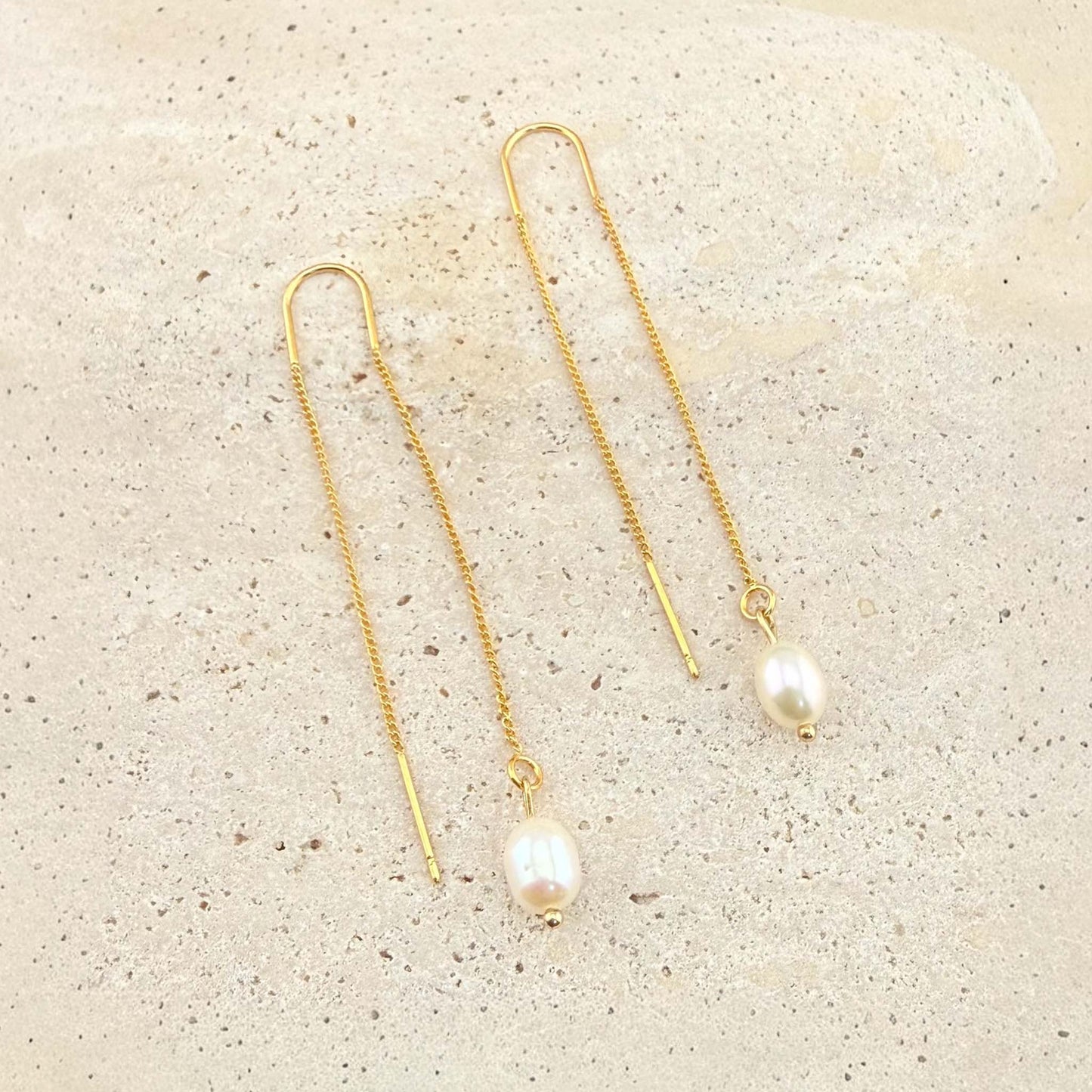 A pair of dainty Aking Sinta gold chain threader earrings, each featuring a classic single white pearl drop at the end of a delicate gold chain. The earrings are laid out elegantly on a light, textured natural stone surface, showcasing their minimalist and timeless appeal