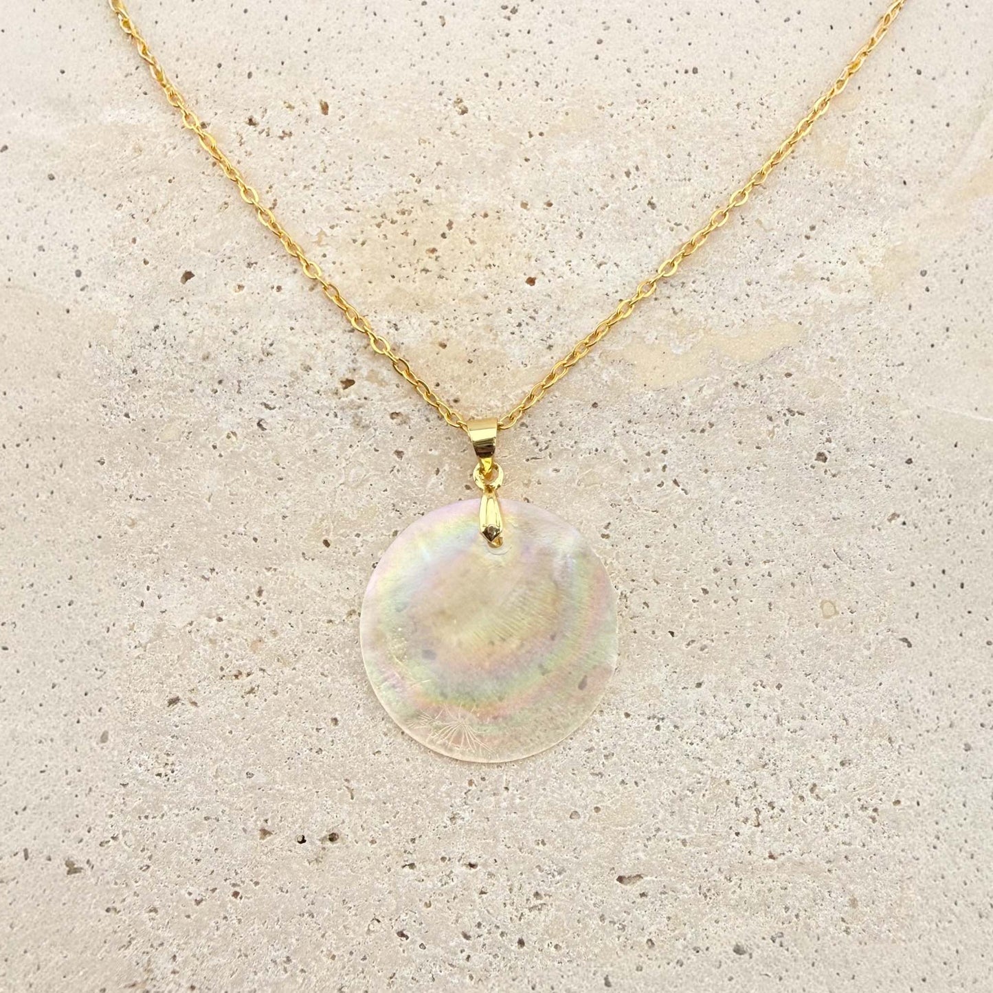 A close-up view of a gold Aking Sinta necklace featuring a large, circular Capiz shell pendant. The iridescent, natural shell disc captures light, displaying beautiful rainbow-like shimmering hues, suspended from a polished gold bail and a delicate gold cable chain on a light, textured natural stone surface