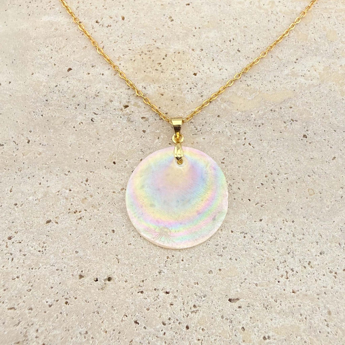 A gold Aking Sinta necklace featuring a large, circular Capiz shell pendant. The iridescent, natural shell disc exhibits beautiful rainbow-like shimmering hues, suspended from a polished gold bail and a delicate gold cable chain on a light, textured natural stone surface