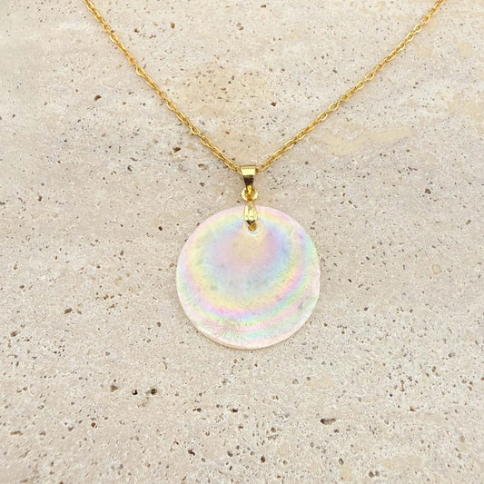 A gold Aking Sinta necklace featuring a large, circular Capiz shell pendant. The iridescent, natural shell disc exhibits beautiful rainbow-like shimmering hues, suspended from a polished gold bail and a delicate gold cable chain on a light, textured natural stone surface