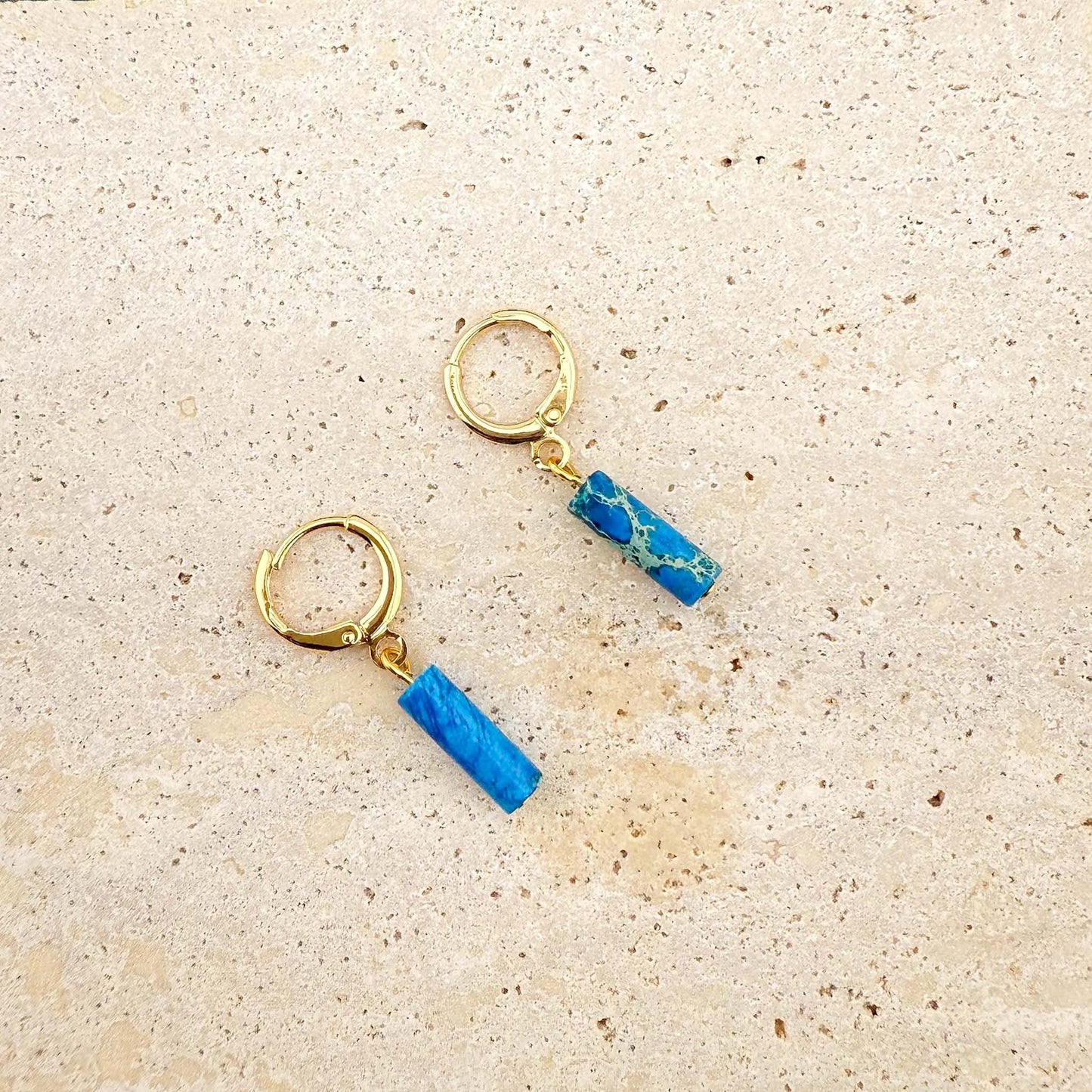 Gold Aking Sinta huggie earrings with circular ocean blue Imperial Jasper stone charms on a white marble background.