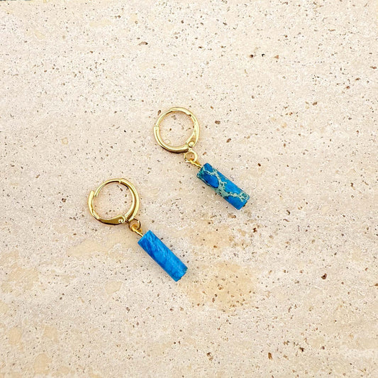 Gold Aking Sinta huggie earrings with circular ocean blue Imperial Jasper stone charms on a white marble background.