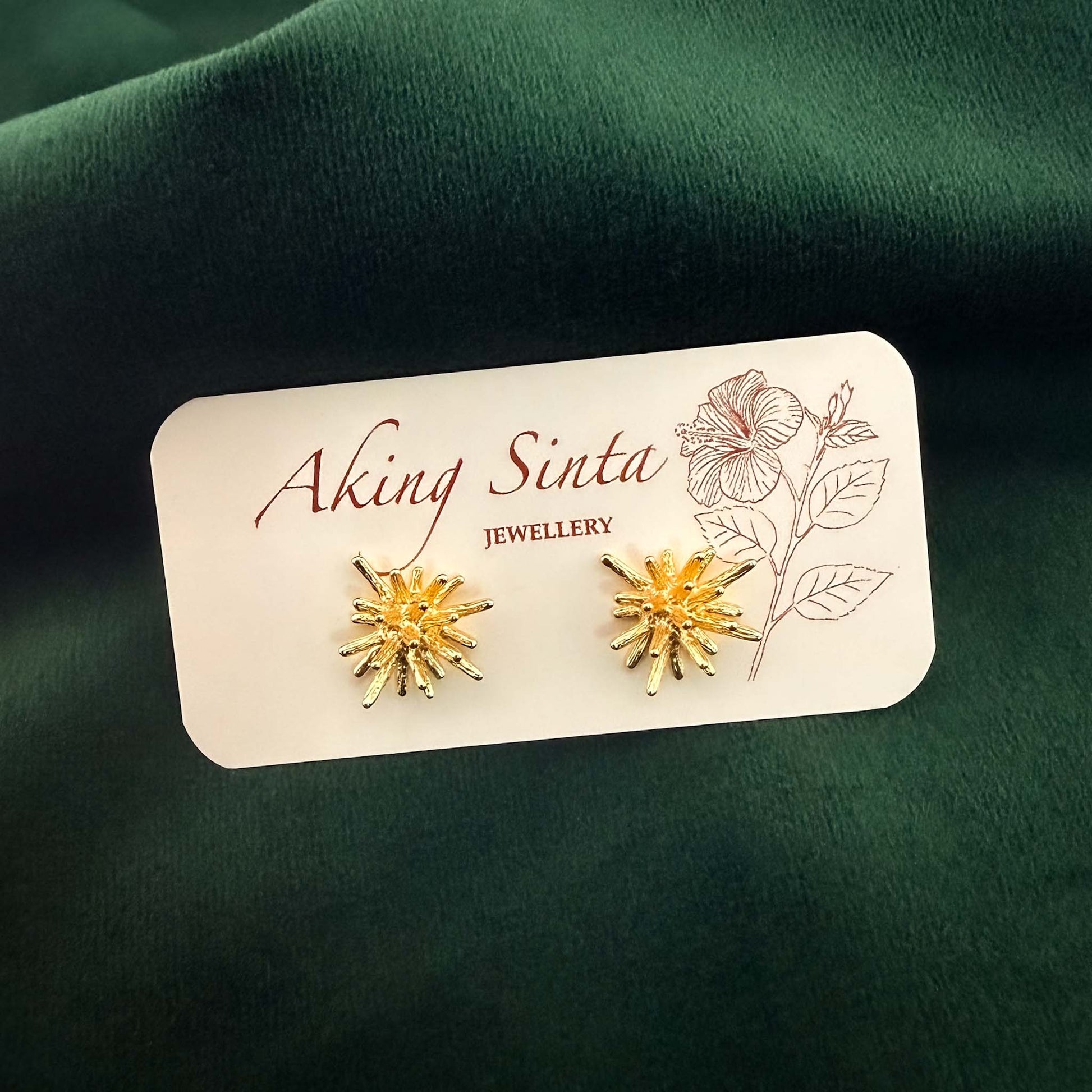 A pair of Aking Sinta gold coral starburst stud earrings pinned to a white jewelry display card featuring the brand name and a pink hibiscus line drawing. The textured, organic-shaped studs are photographed without their drop backs against a rich, dark emerald green velvet fabric background