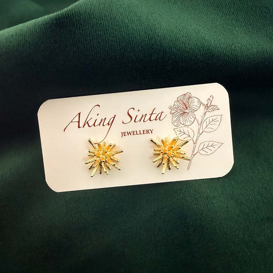 A pair of Aking Sinta gold coral starburst stud earrings pinned to a white jewelry display card featuring the brand name and a pink hibiscus line drawing. The textured, organic-shaped studs are photographed without their drop backs against a rich, dark emerald green velvet fabric background