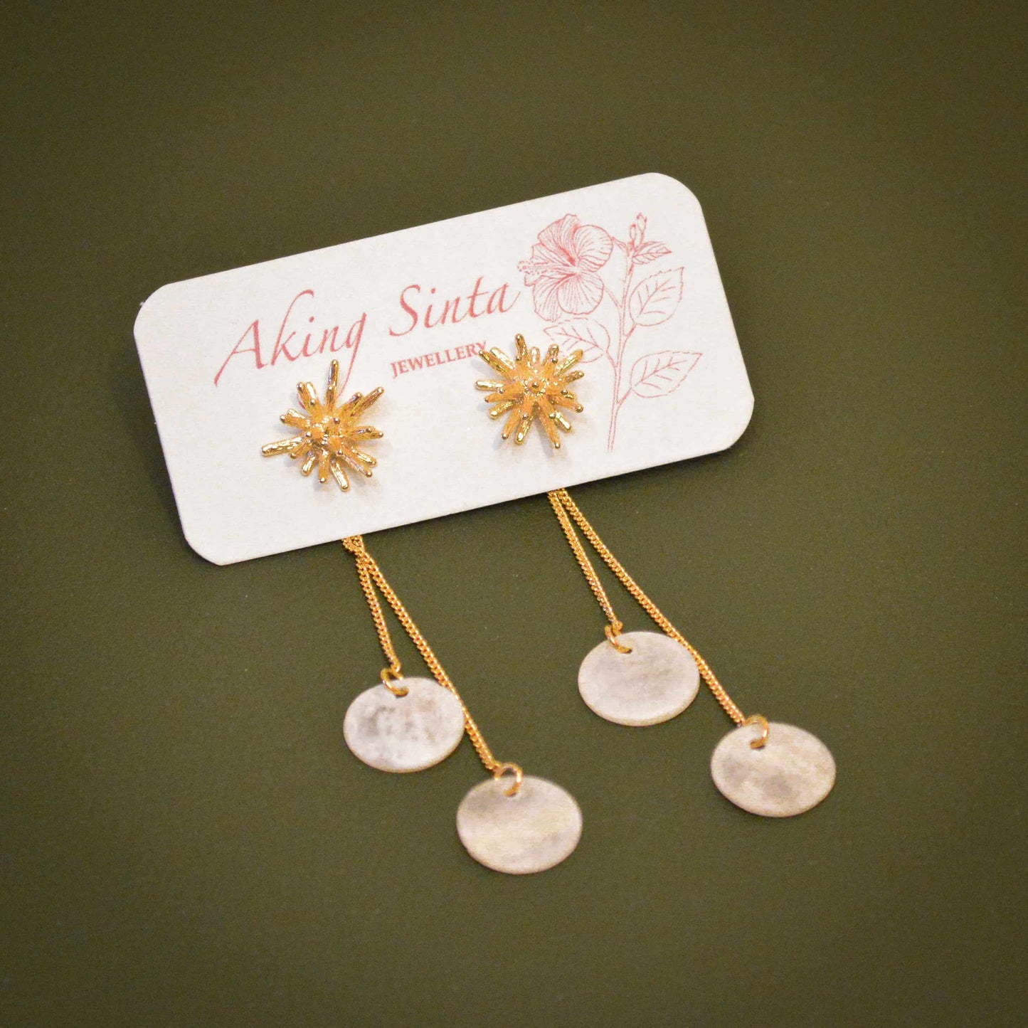 A pair of Aking Sinta 2-in-1 gold stud earrings displayed on a white branded jewelry card with a pink hibiscus illustration. The earrings feature textured gold coral starburst studs paired with removable double chain drop backs that suspend translucent, round white Capiz shell discs against an olive green background.