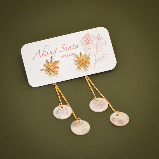 A pair of Aking Sinta 2-in-1 gold stud earrings displayed on a white branded jewelry card with a pink hibiscus illustration. The earrings feature textured gold coral starburst studs paired with removable double chain drop backs that suspend translucent, round white Capiz shell discs against an olive green background.