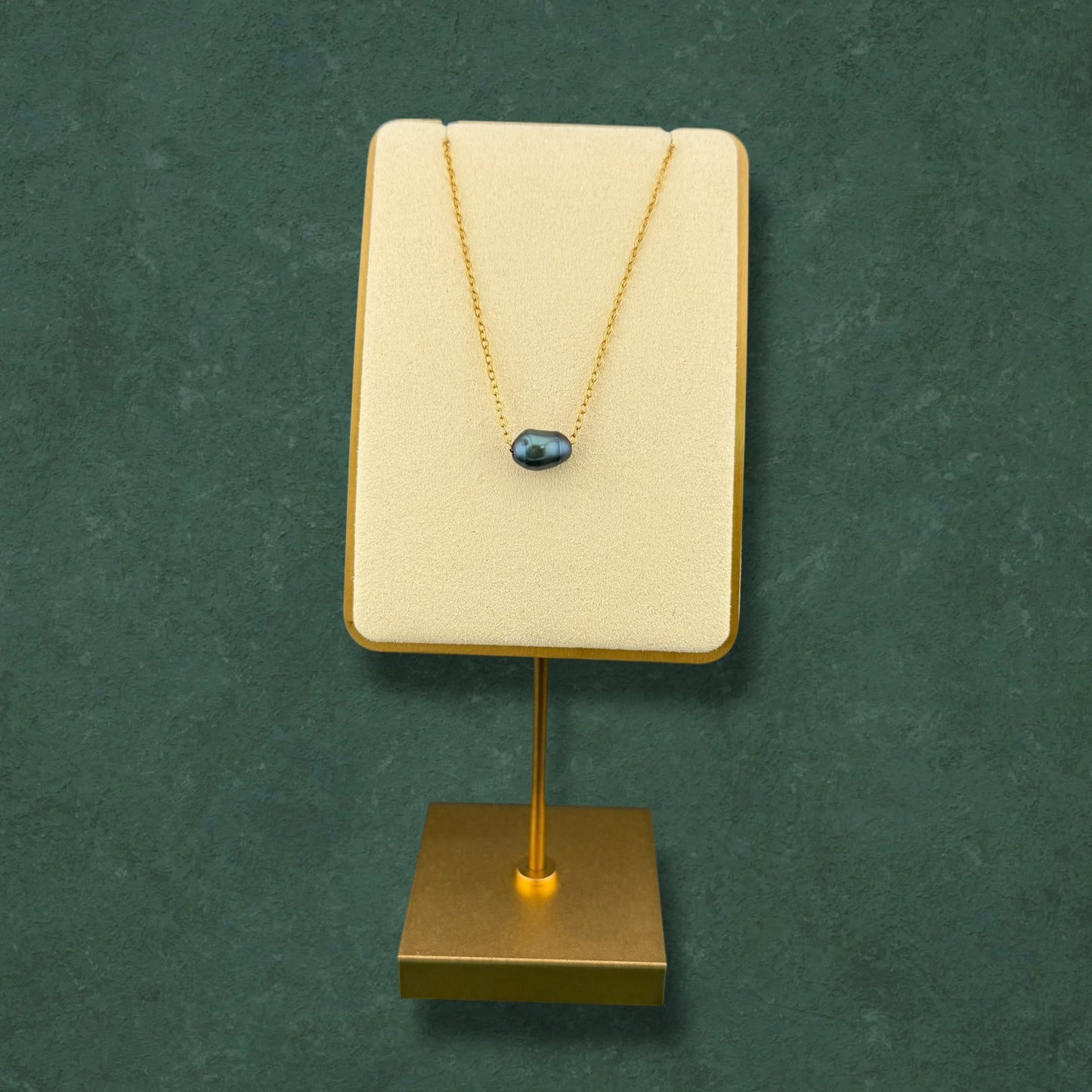 A minimalist Aking Sinta dainty gold chain necklace featuring a single, iridescent dark pearl pendant. The necklace is elegantly showcased on a cream-colored velvet stand with a gold base, set against a rich, dark green textured background, highlighting its sophisticated and refined design