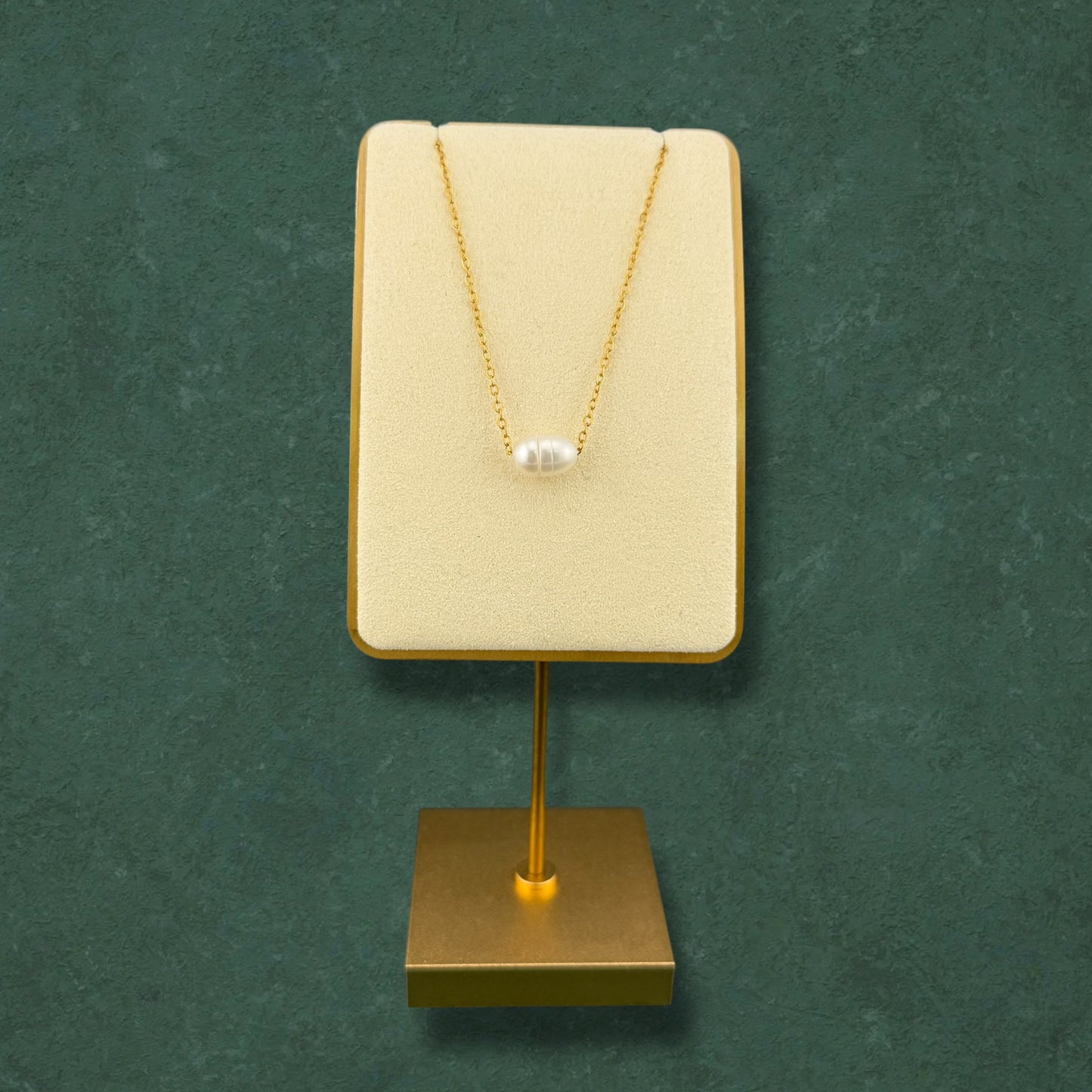A minimalist Aking Sinta dainty gold chain necklace featuring a single, lustrous white pearl pendant. The necklace is elegantly displayed on a cream-colored velvet stand with a gold base, set against a rich, dark green textured background, emphasizing its sophisticated and refined design