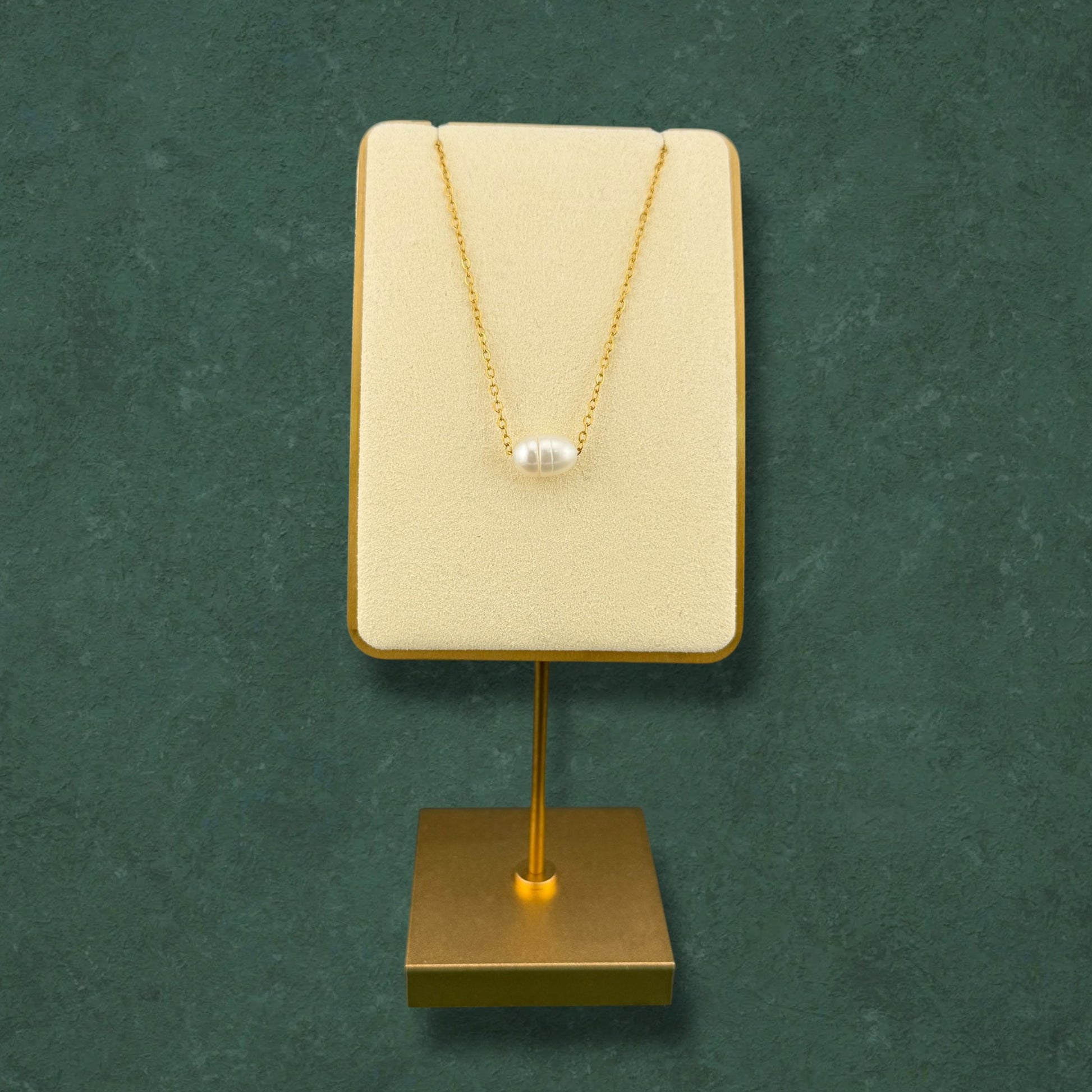 A minimalist Aking Sinta dainty gold chain necklace featuring a single, lustrous white pearl pendant. The necklace is elegantly displayed on a cream-colored velvet stand with a gold base, set against a rich, dark green textured background, emphasizing its sophisticated and refined design
