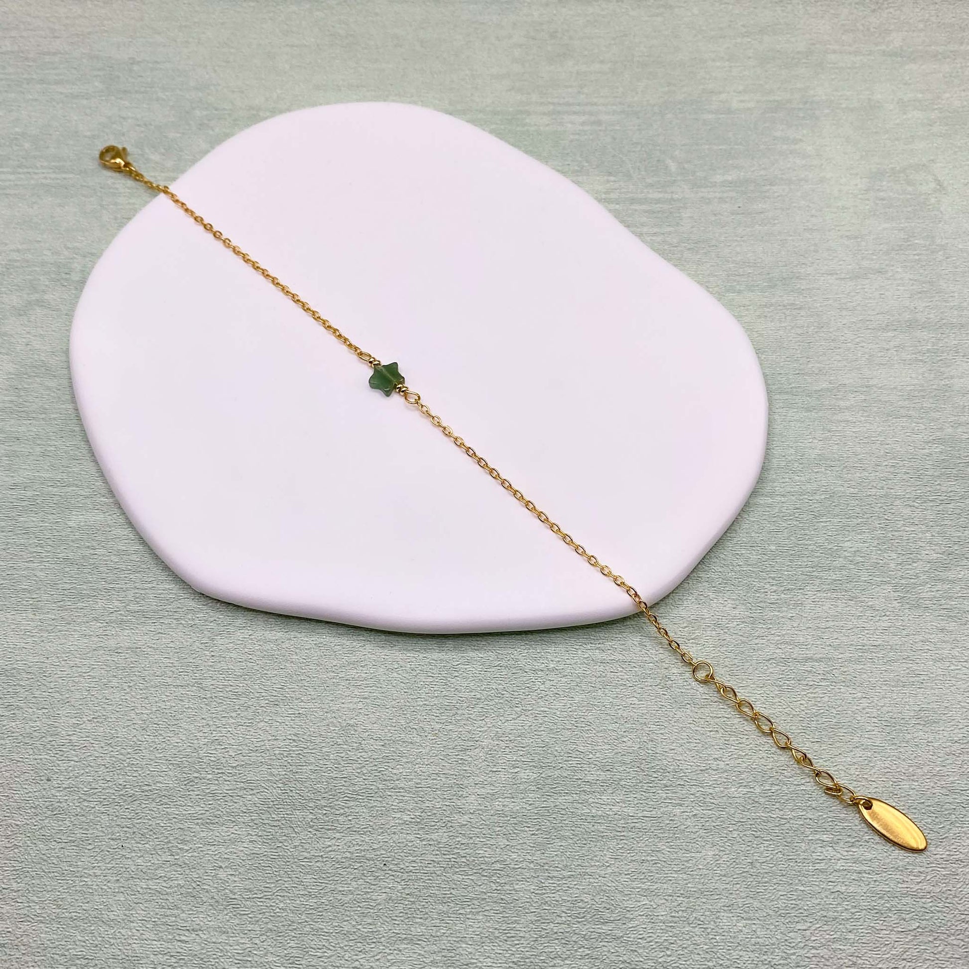 An Aking Sinta product photograph showing the dainty green aventurine star bracelet laid out diagonally across an asymmetrical white display dish over a light sage green background. The delicate gold chain bracelet features a small star-shaped green aventurine gemstone charm at the center, a lobster clasp closure on one end, and an adjustable extender chain ending in an oval gold brand tag on the other