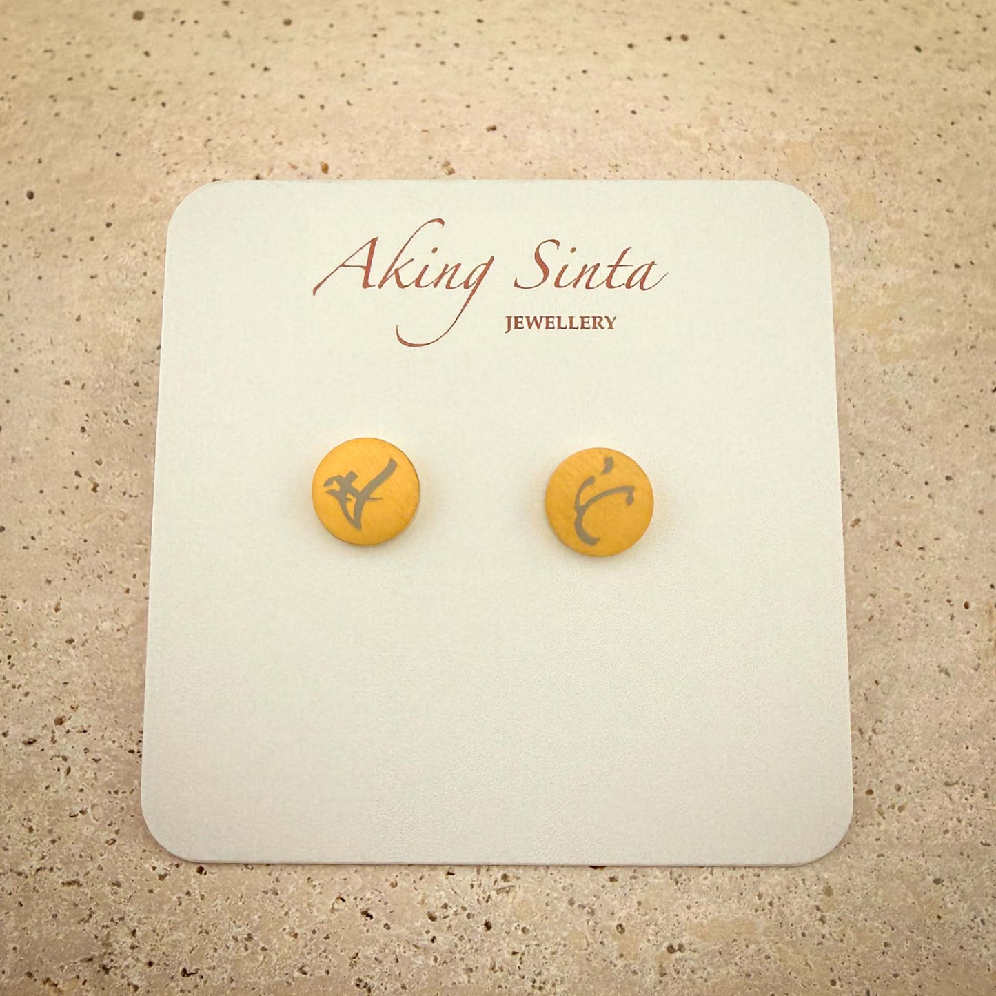 A pair of gold dainty stud earrings from Aking Sinta Jewellery featuring bespoke engravings. The small circular gold studs are etched with ancient Filipino Baybayin script and presented on a white branded display card, set against a light-colored natural stone background