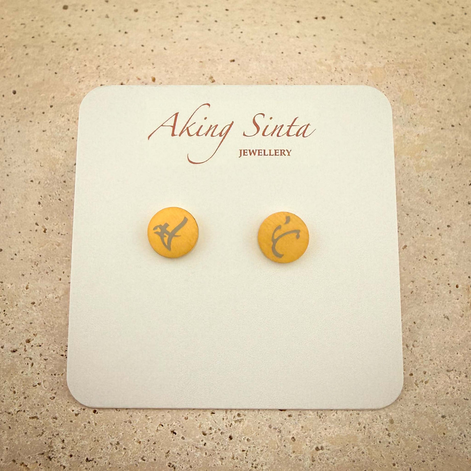 A pair of gold dainty stud earrings from Aking Sinta Jewellery featuring bespoke engravings. The small circular gold studs are etched with ancient Filipino Baybayin script and presented on a white branded display card, set against a light-colored natural stone background