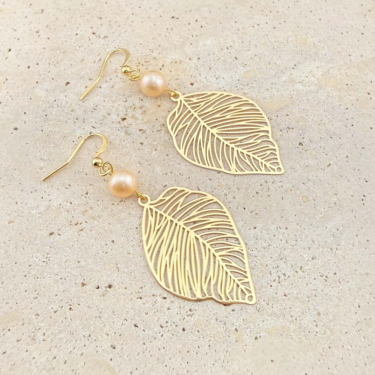 A pair of Aking Sinta gold drop earrings featuring intricate filigree leaf charms and round peach freshwater pearls. The nature-inspired jewelry is displayed on a light-colored natural stone background.