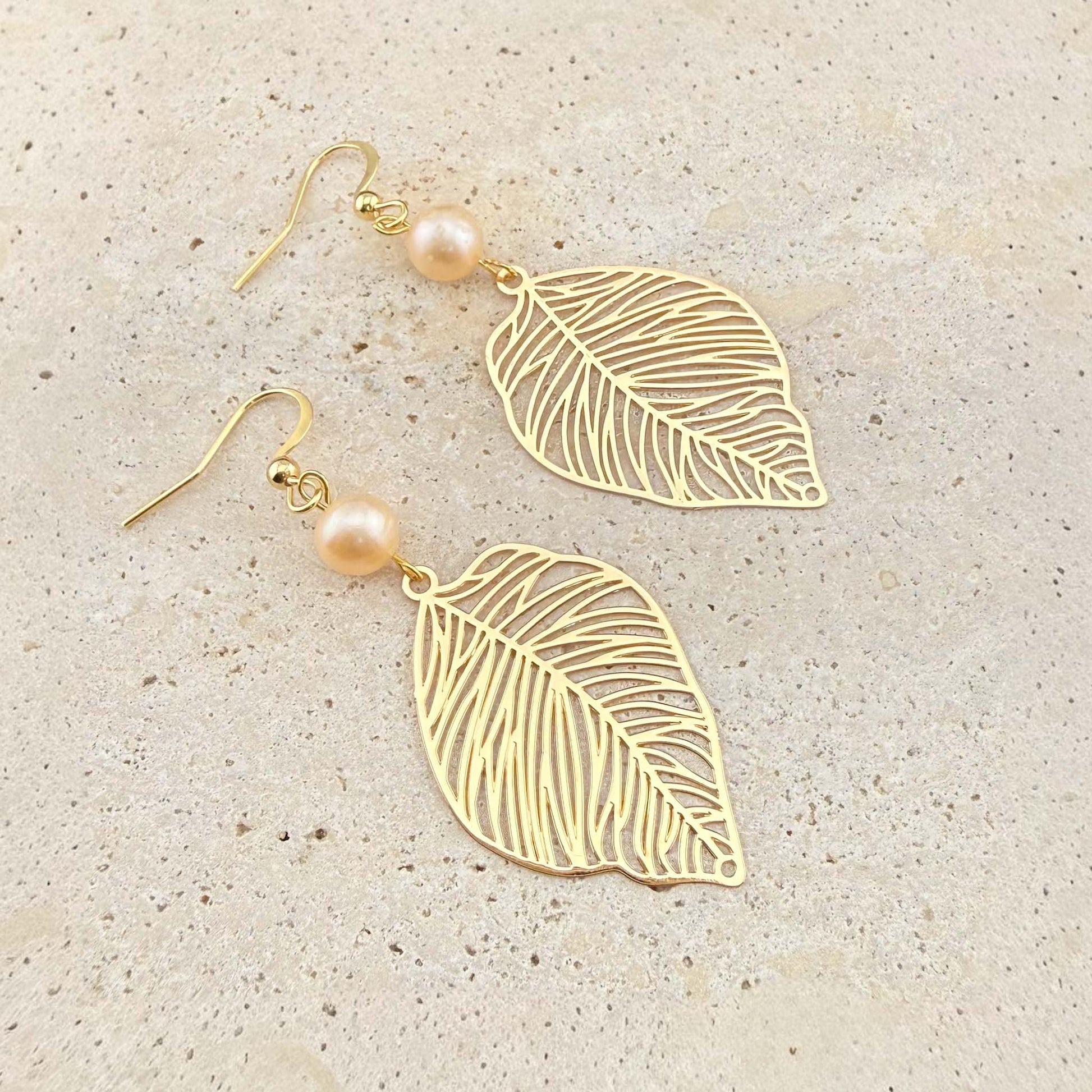 A pair of Aking Sinta gold drop earrings featuring intricate filigree leaf charms and round peach freshwater pearls. The nature-inspired jewelry is displayed on a light-colored natural stone background.