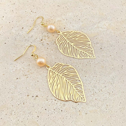 A pair of Aking Sinta gold drop earrings featuring intricate filigree leaf charms and round peach freshwater pearls. The nature-inspired jewelry is displayed on a light-colored natural stone background.