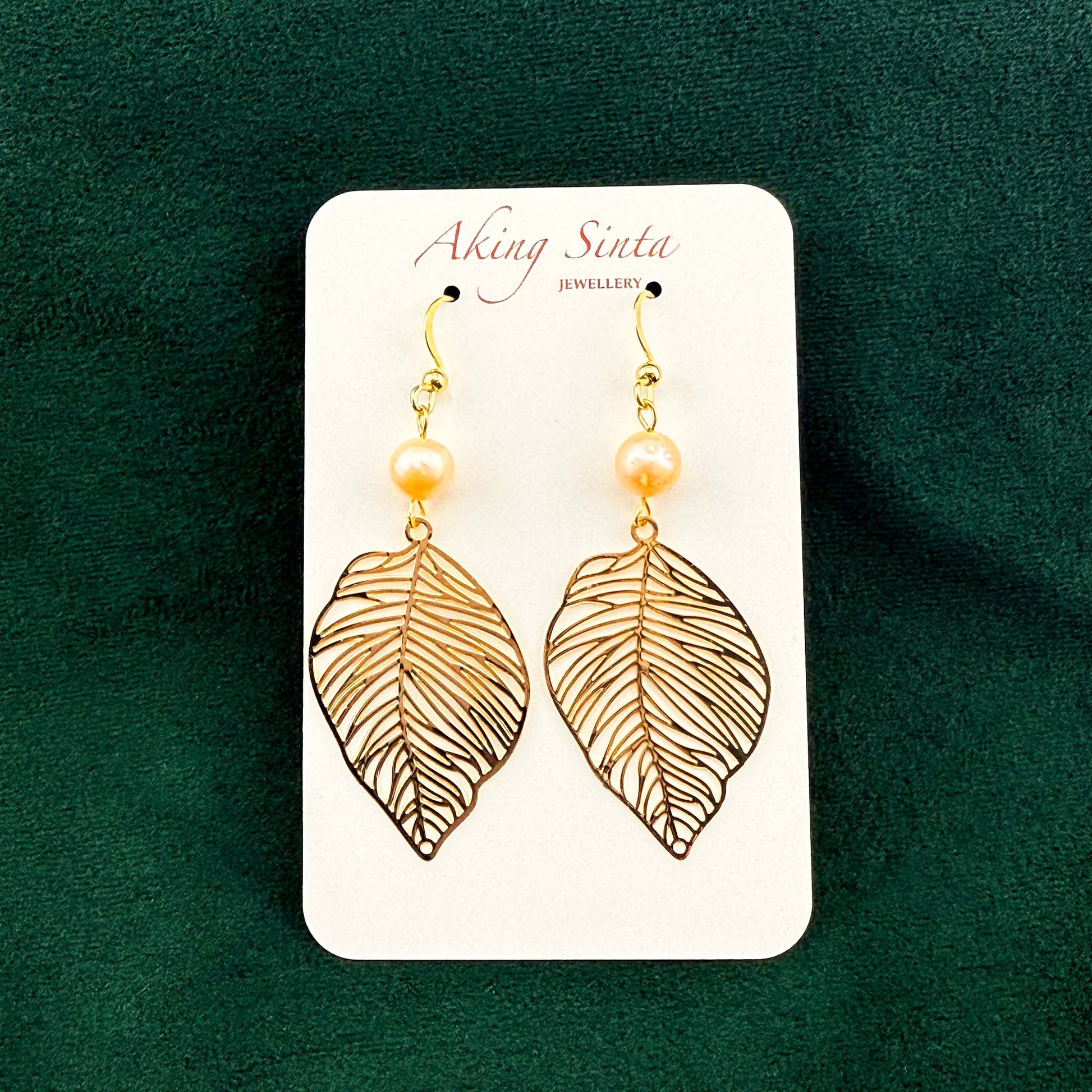 A pair of Aking Sinta gold filigree leaf and peach pearl drop earrings on a white branded display card. The delicate, nature-inspired earrings are set against a rich dark green velvet background.