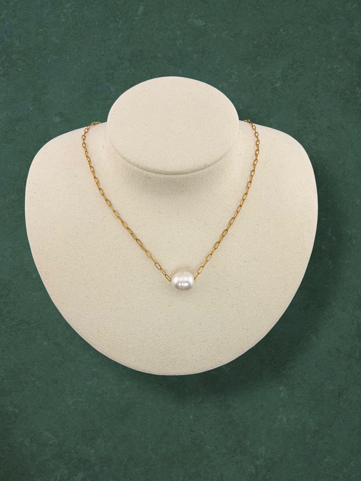 Aking Sinta gold paperclip chain necklace featuring a single floating white baroque pearl. Minimalist jewelry shown on a white velvet bust display against a textured green background.