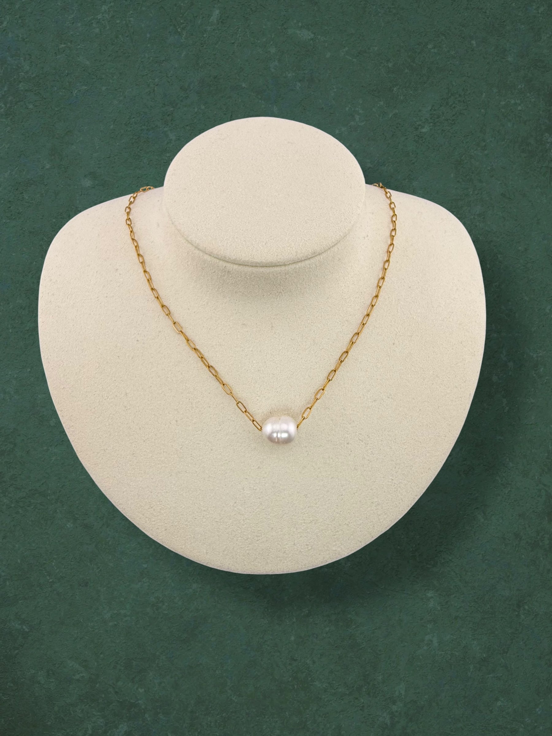 Aking Sinta gold paperclip chain necklace featuring a single floating white baroque pearl. Minimalist jewelry shown on a white velvet bust display against a textured green background.