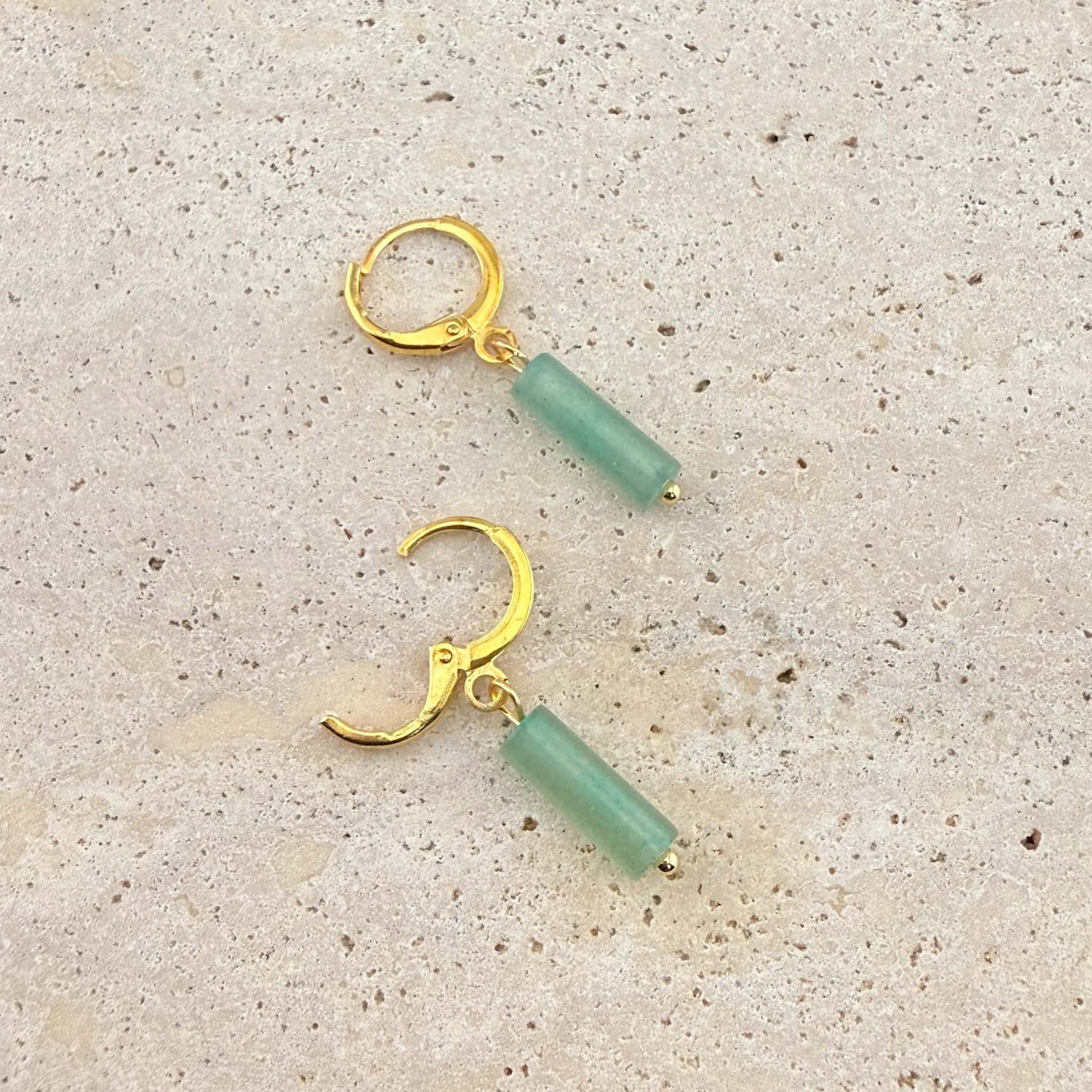 A pair of Aking Sinta gold huggie hoop earrings with green aventurine column drops. One earring is open to show the clasp mechanism, presented on a natural light-colored stone background