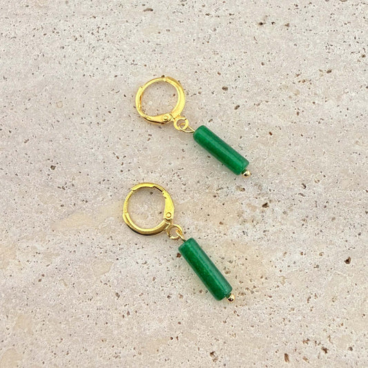 A pair of Aking Sinta gold huggie hoop earrings featuring vibrant green jade column drop pendants. These polished gemstone earrings are displayed on a natural, light-colored stone surface