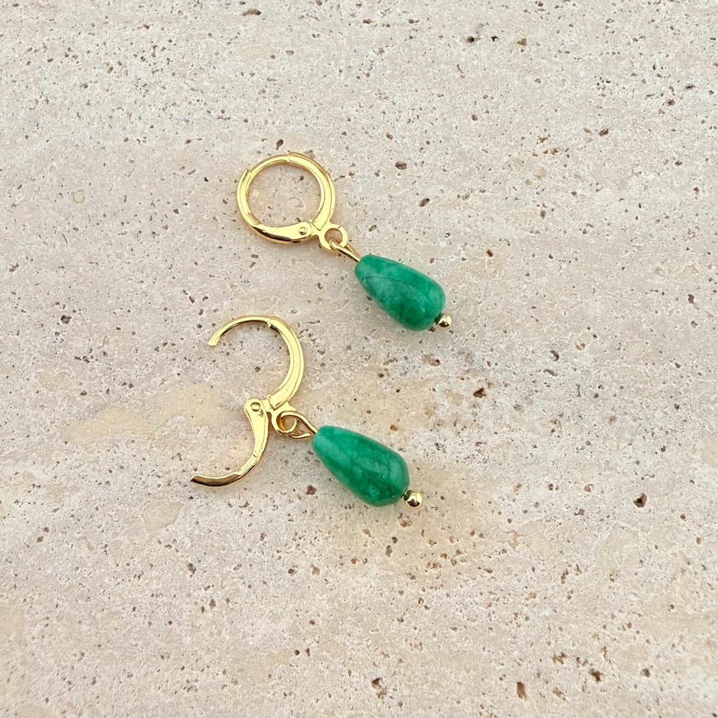 A pair of Aking Sinta gold huggie earrings with polished green jade teardrop charms. One earring is shown open to highlight the easy-to-use clasp on a natural stone surface