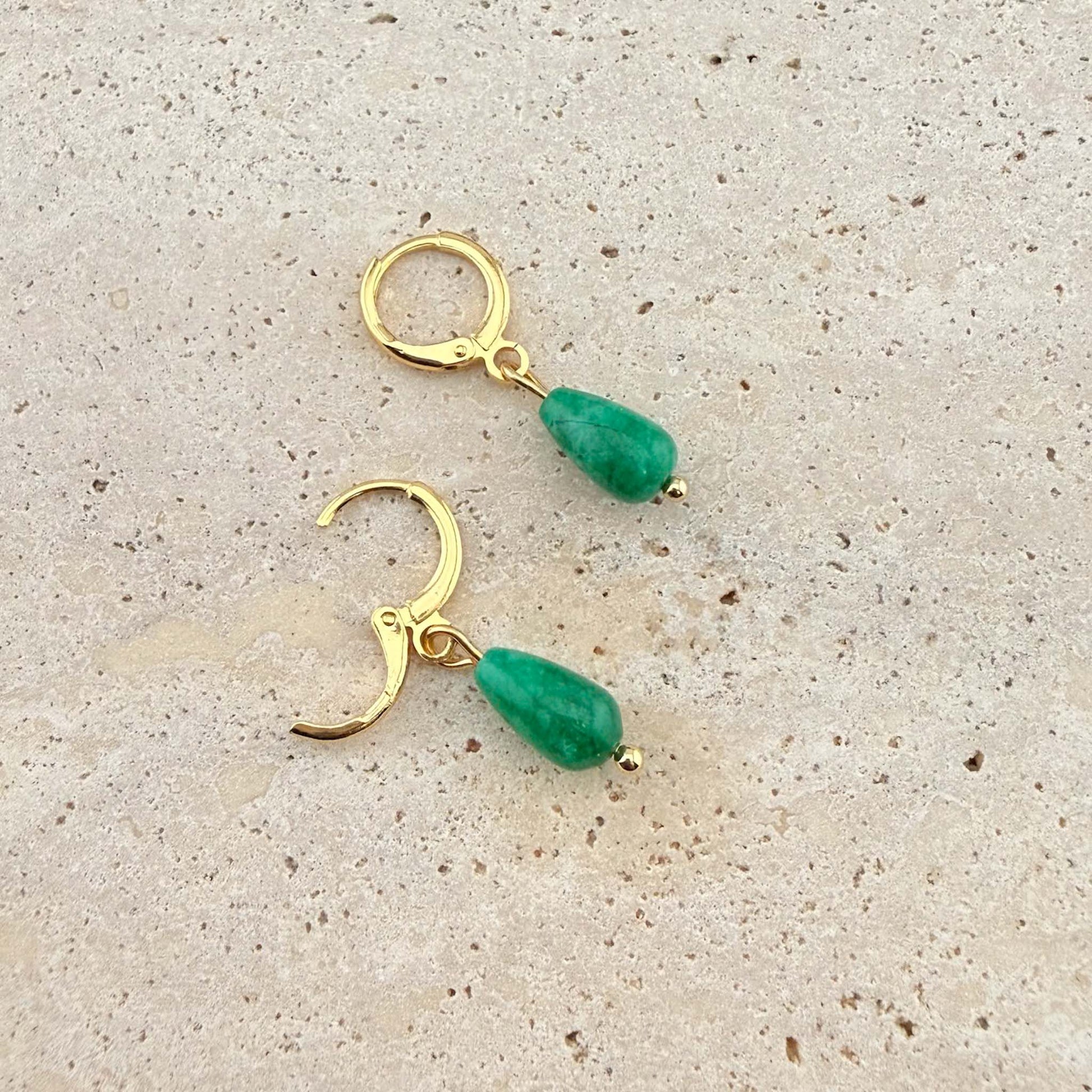 A pair of Aking Sinta gold huggie earrings with polished green jade teardrop charms. One earring is shown open to highlight the easy-to-use clasp on a natural stone surface