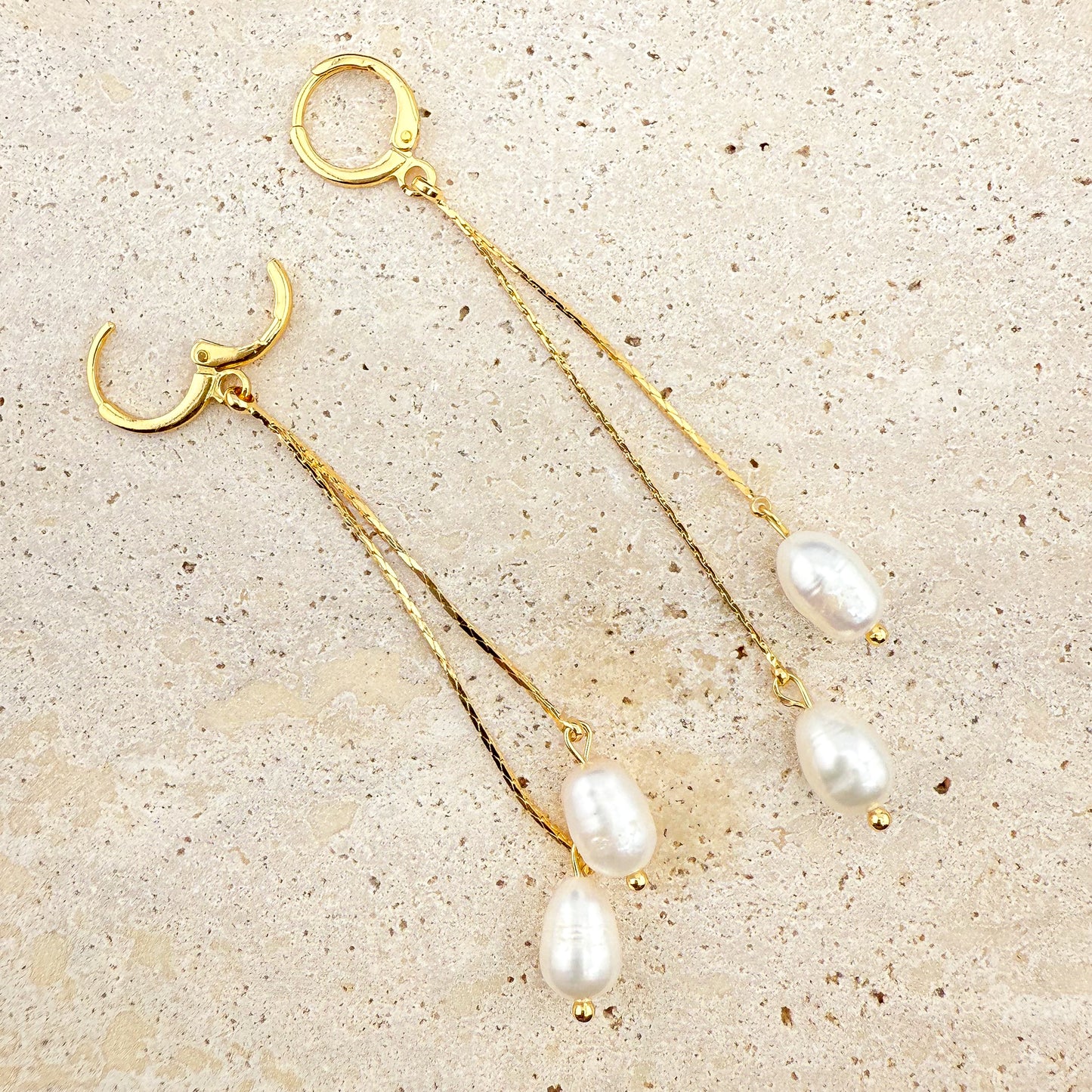 A pair of Aking Sinta gold earrings featuring a secure hinge-clasp design. Each earring has two delicate gold chains of varying lengths, each ending in a lustrous white pearl drop. The earrings are artfully arranged on a light, textured natural stone surface, highlighting the functional hinge detail and the elegant, layered aesthetic