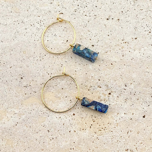 A pair of Aking Sinta gold hoop earrings featuring vibrant blue imperial jasper cuboid gemstone pendants. The minimalist gold wire hoops and unique marbled stone drops are displayed on a light-colored natural stone surface.