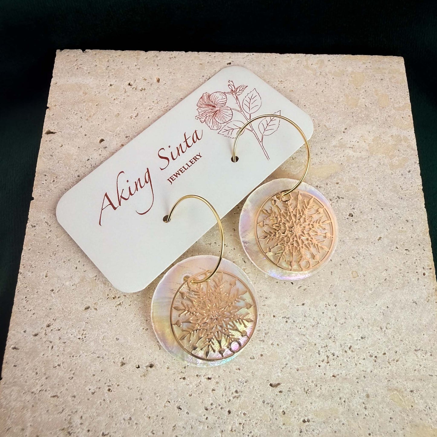 A pair of Aking Sinta gold hoop earrings featuring iridescent round Capiz shell discs and intricate gold snowflake charms. The earrings are displayed on a branded white jewelry card with a hibiscus illustration, resting on a light, textured natural stone block against a dark background