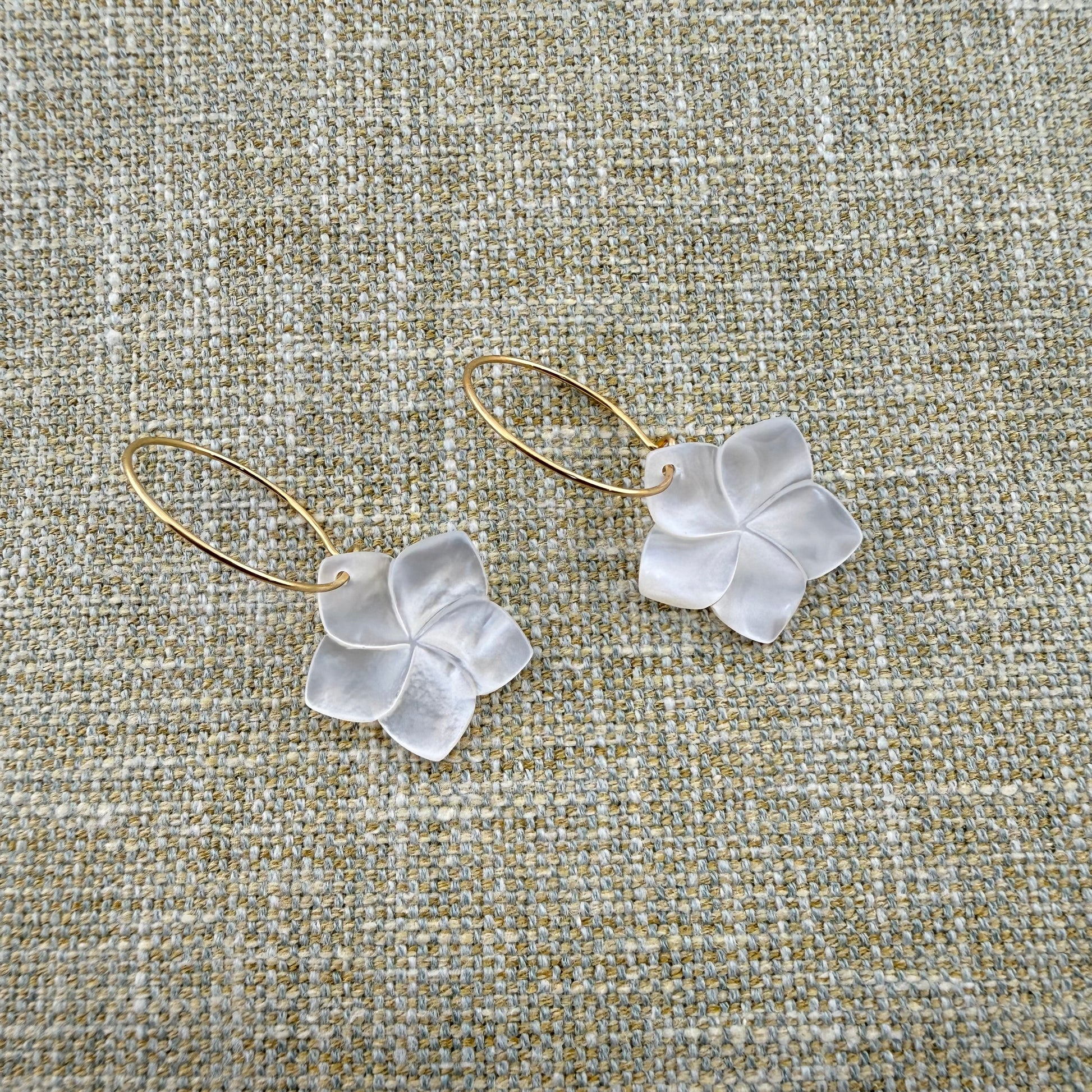 A pair of Aking Sinta gold hoop earrings featuring hand-carved Mother of Pearl Kalachuchi (Plumeria) flower drops. The iridescent white shell petals catch the light, showcasing their natural luster. The earrings are laid out on a neutral, woven linen fabric background, highlighting the delicate tropical design
