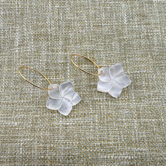 A pair of Aking Sinta gold hoop earrings featuring hand-carved Mother of Pearl Kalachuchi (Plumeria) flower drops. The iridescent white shell petals catch the light, showcasing their natural luster. The earrings are laid out on a neutral, woven linen fabric background, highlighting the delicate tropical design