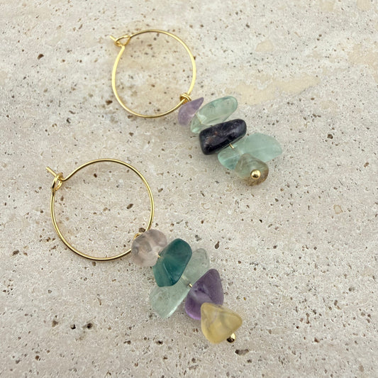 A pair of Aking Sinta gold hoop earrings featuring dangling, multi-colored raw fluorite nugget chips. The natural gemstone chips display a vibrant mix of green, purple, and yellow hues, elegantly displayed on a light, textured natural stone background