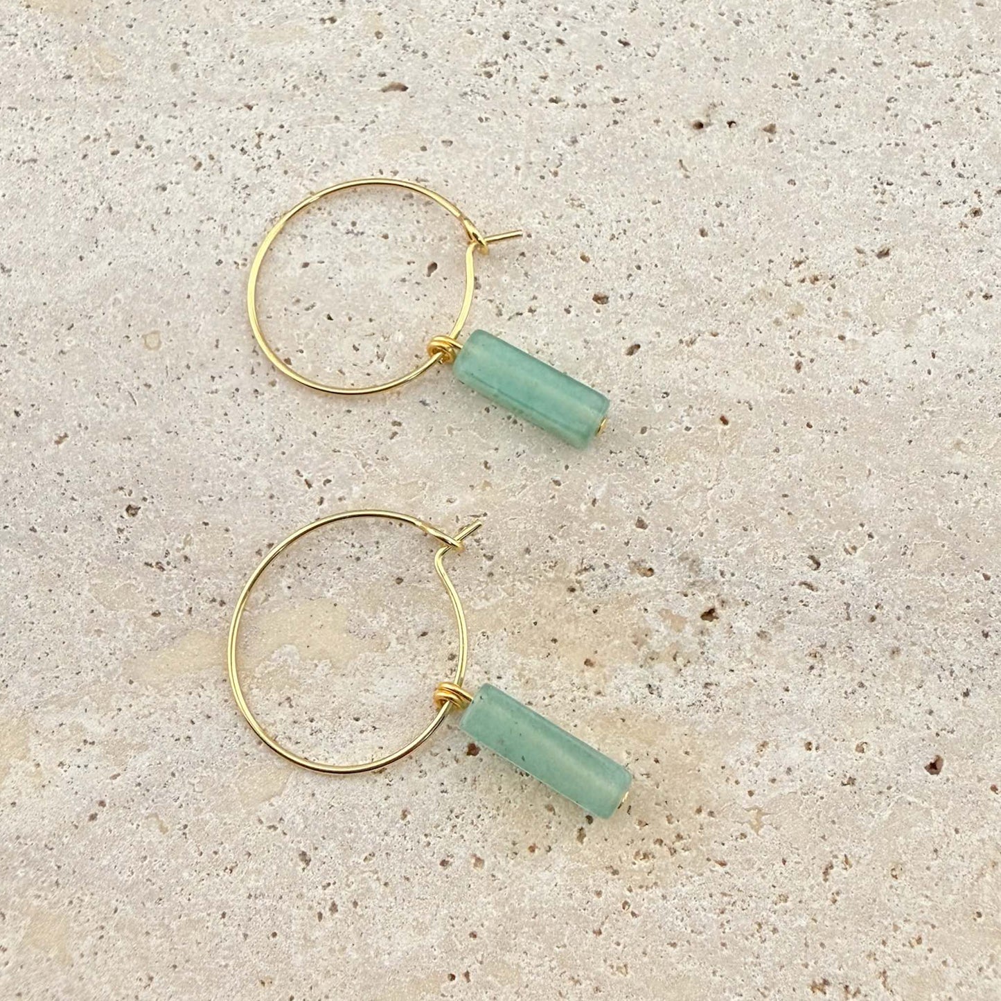 A pair of Aking Sinta gold hoop earrings featuring light green aventurine cuboid-shaped gemstone pendants. The minimalist wire hoops and natural stone drops are displayed on a light-colored natural stone surface.
