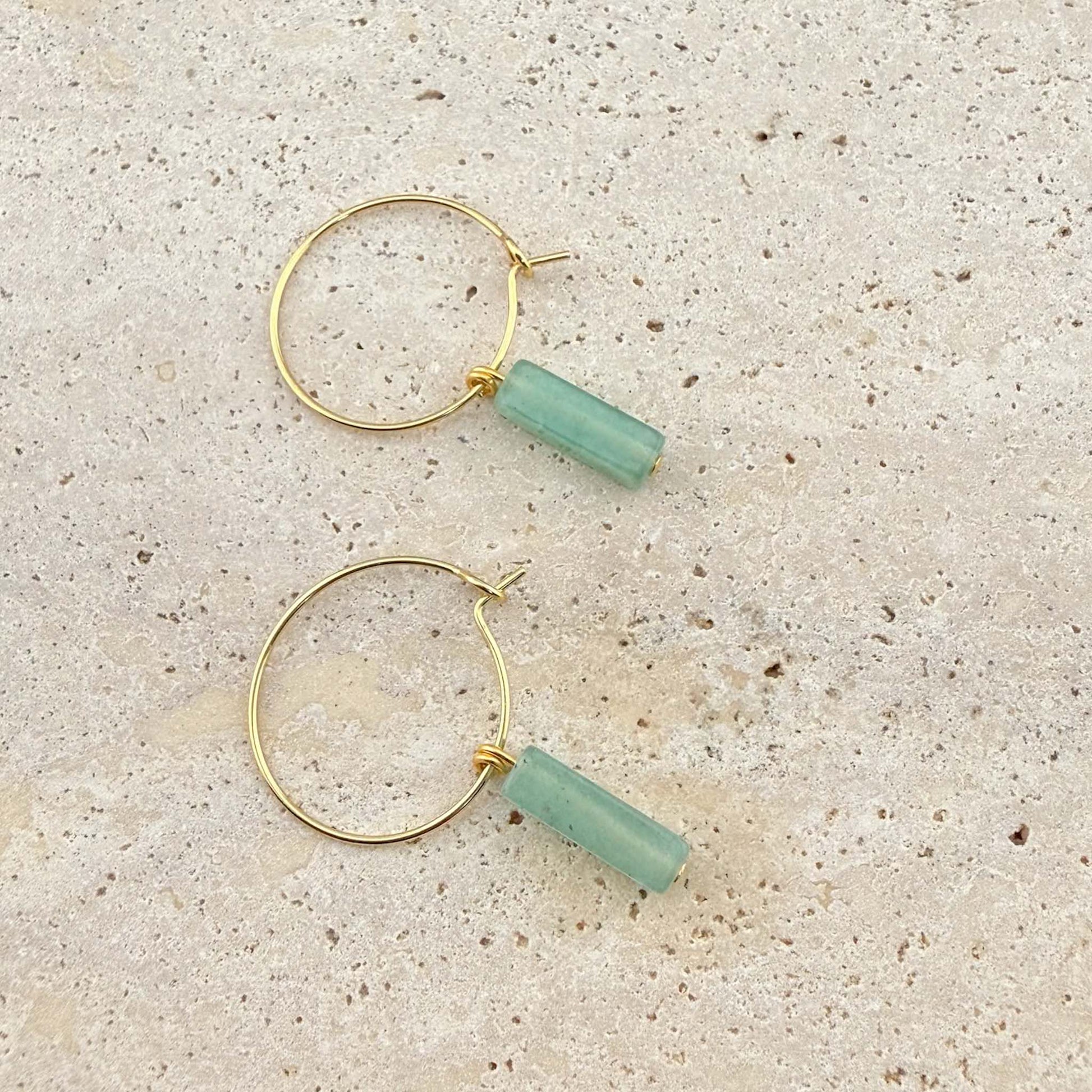 A pair of Aking Sinta gold hoop earrings featuring light green aventurine cuboid-shaped gemstone pendants. The minimalist wire hoops and natural stone drops are displayed on a light-colored natural stone surface.