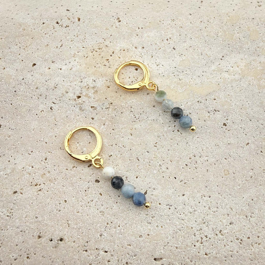 Pair of Aking Sinta gold huggie earrings featuring round, translucent Aquamarine beads on a white marble texture.