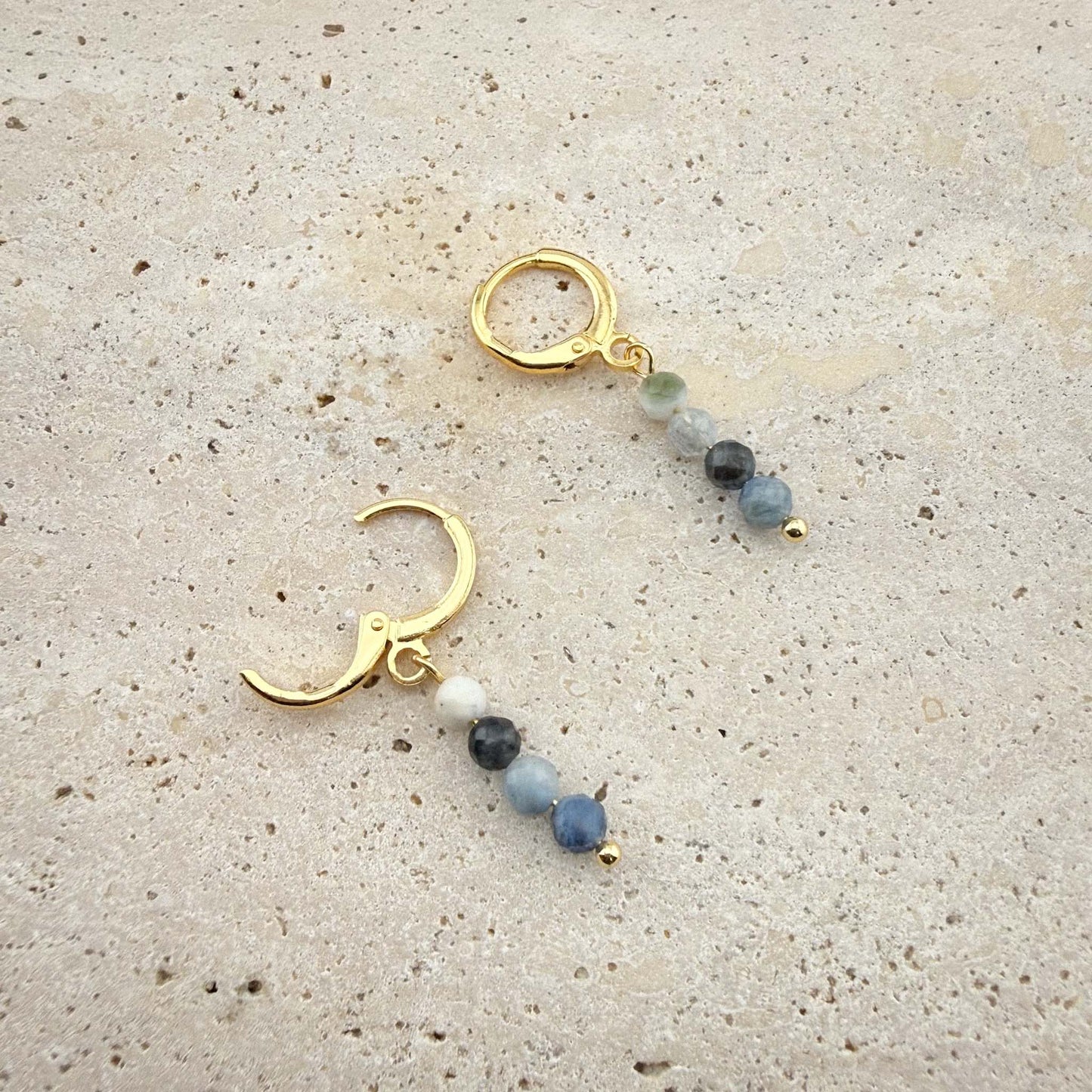 Close-up of gold Aking Sinta huggie earrings with round Aquamarine beads, showing one hoop closed and one hoop with the hinge clasp open on marble.