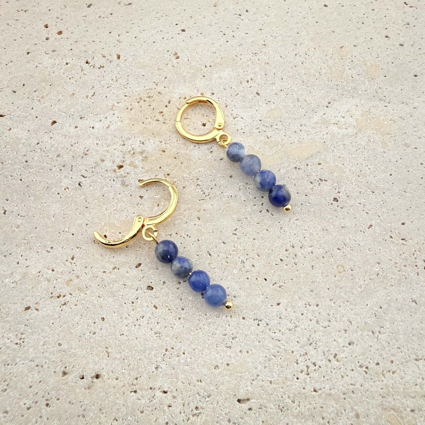 Close-up of gold Aking Sinta huggie earrings with three matte blue beads, showing one hoop closed and one hoop with the hinge clasp open on marble.