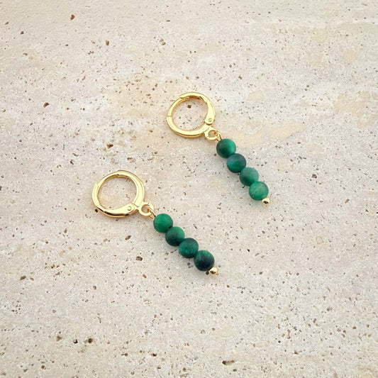 Pair of gold Aking Sinta huggie earrings with three chatoyant Green Tiger Eye beads on a white marble surface.