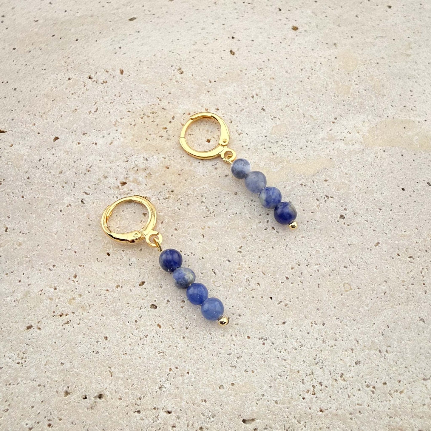 Pair of Aking Sinta gold huggie earrings with three matte blue beads, both hoops securely closed on a white marble background.