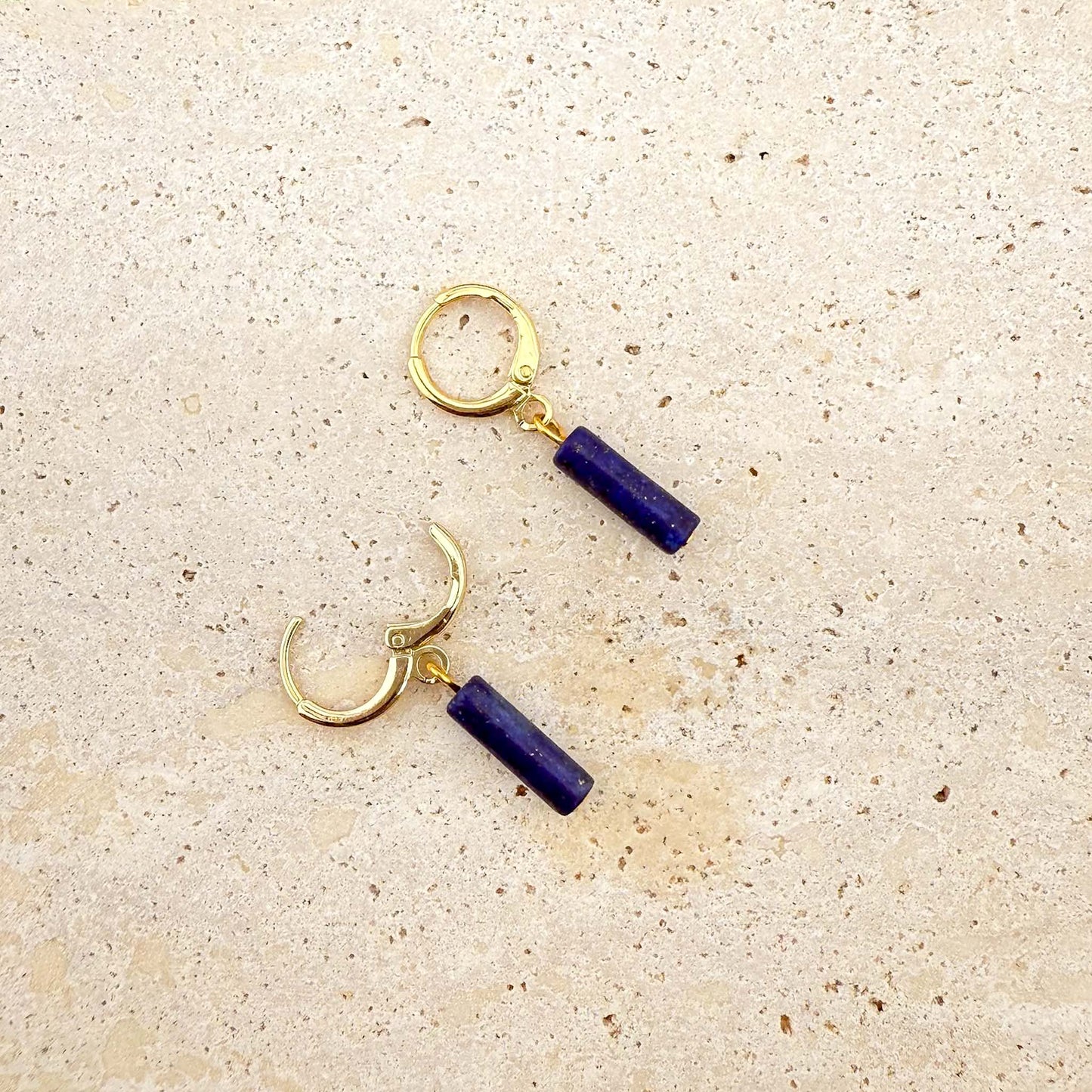 A pair of gold Aking Sinta huggie hoop earrings featuring deep blue lapis lazuli column charms. One hoop is shown with the clasp open, showcasing the hinge design. The natural gemstone pendants feature a rich blue color and hang from polished gold-toned hoops on a light natural stone surface