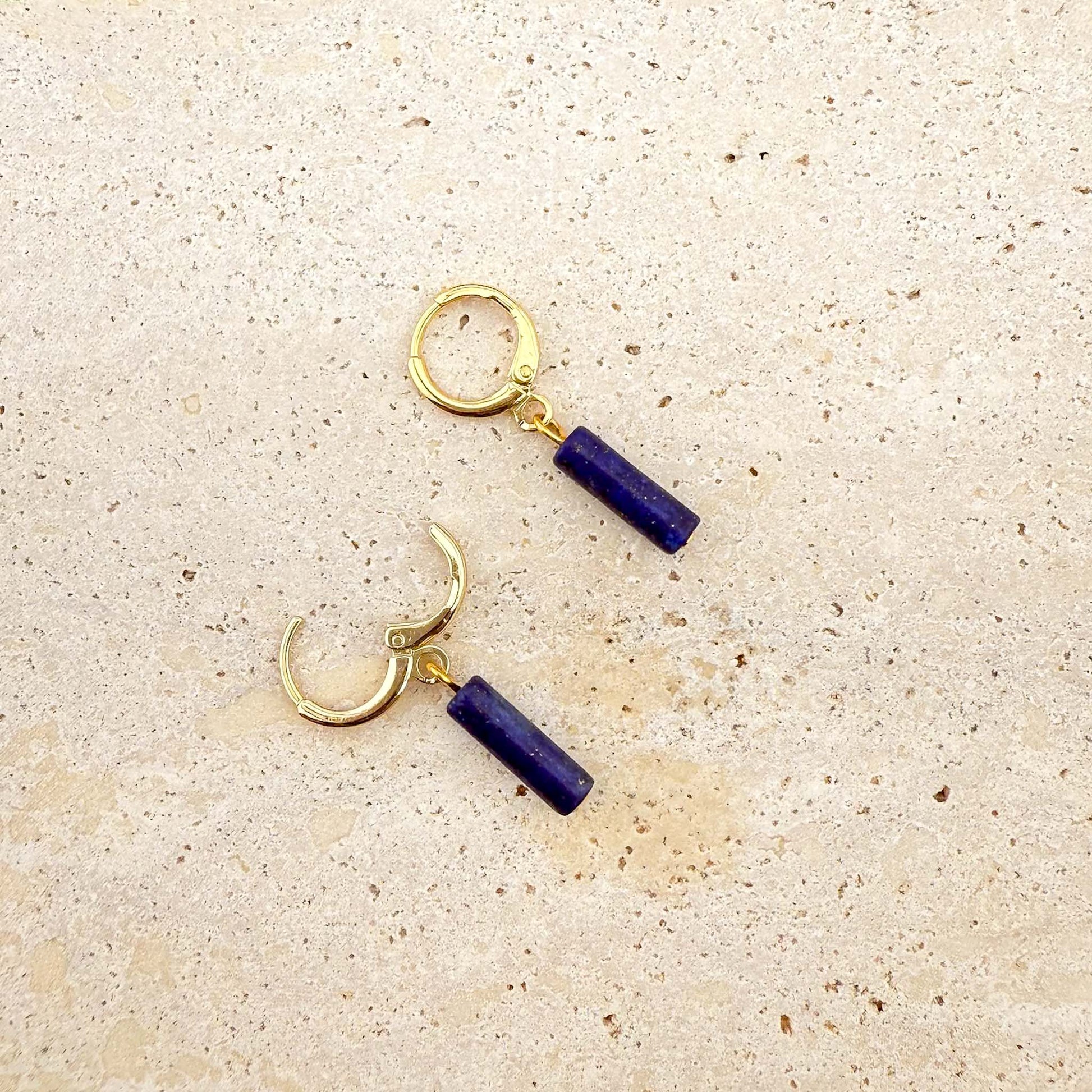A pair of gold Aking Sinta huggie hoop earrings featuring deep blue lapis lazuli column charms. One hoop is shown with the clasp open, showcasing the hinge design. The natural gemstone pendants feature a rich blue color and hang from polished gold-toned hoops on a light natural stone surface